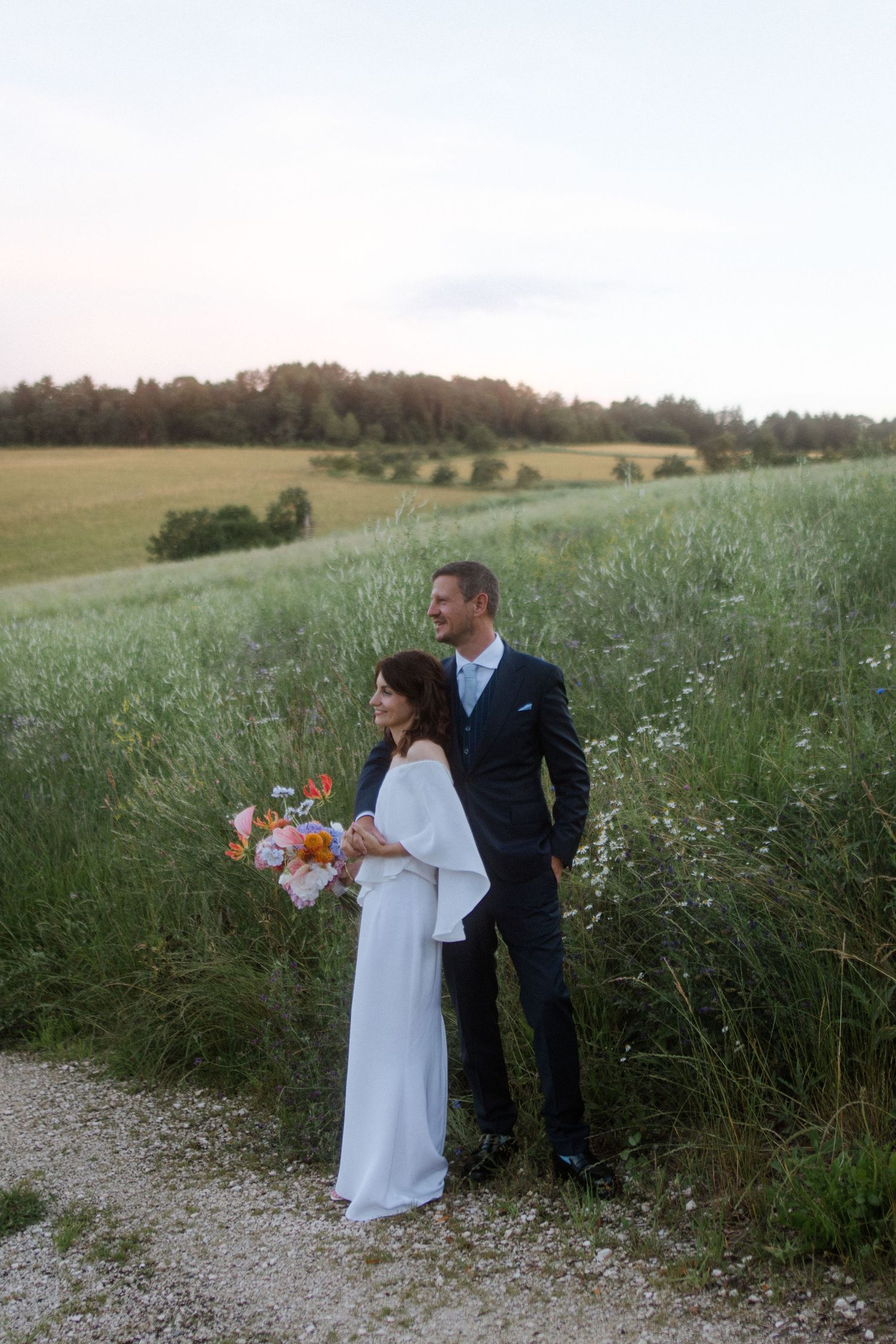 A couple stands together on a grassy path with rolling hills in the background.