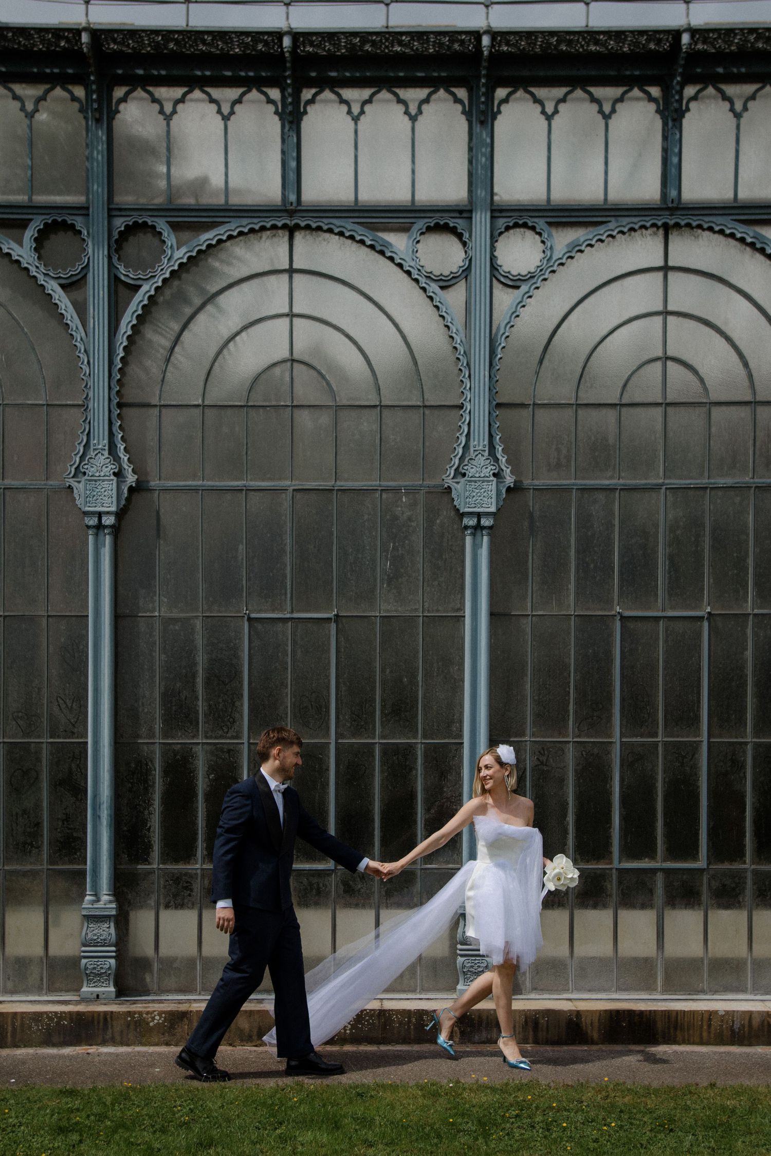 A couple poses in front of a vintage glass conservatory with ornate metalwork details.