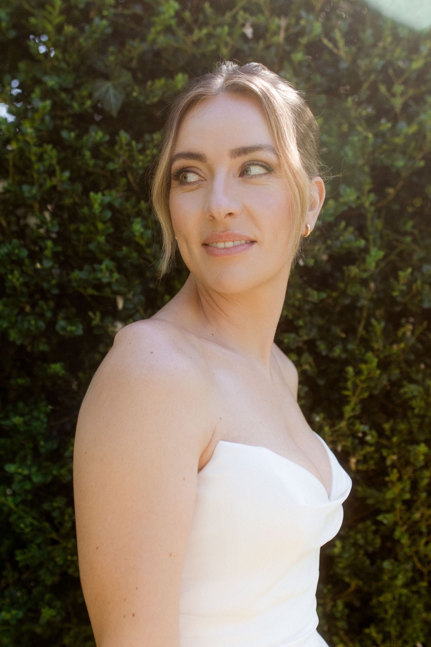 A bride in a strapless white gown poses outdoors against lush green hedges in warm sunlight.