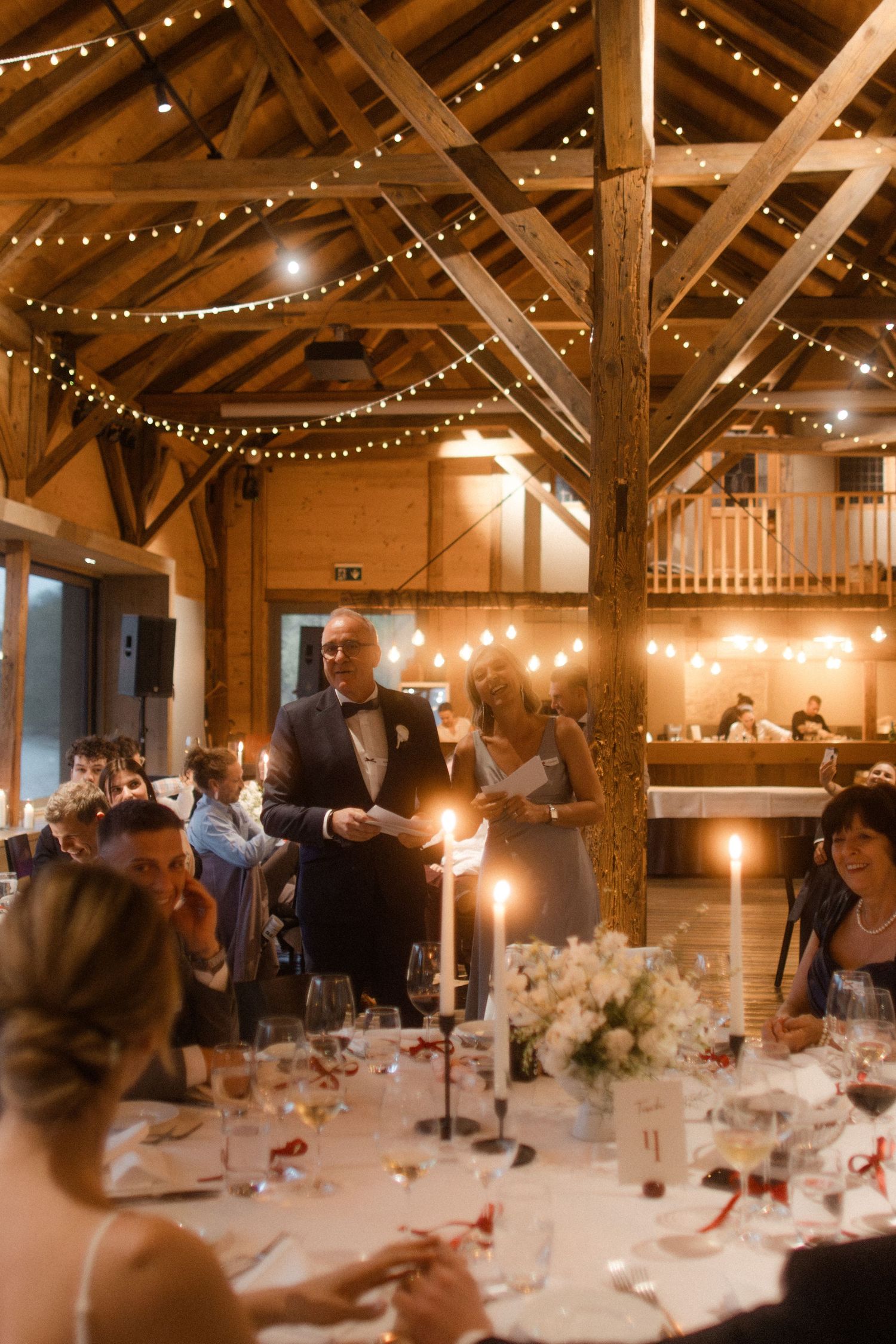 Romantic barn wedding reception with string lights, wooden beams, candlelit tables, and a speaker giving a toast to guests.
