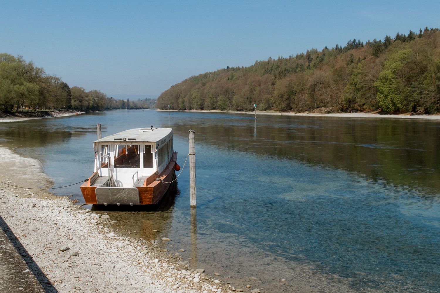 A small wooden boat moored on a calm river shore surrounded by lush green trees under a clear blue sky.