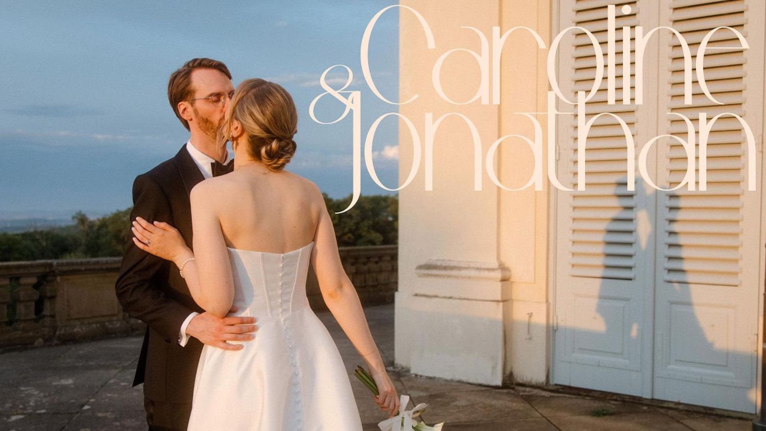 Romantic couple in formal wedding attire embrace and dance together at sunset against white shuttered windows.