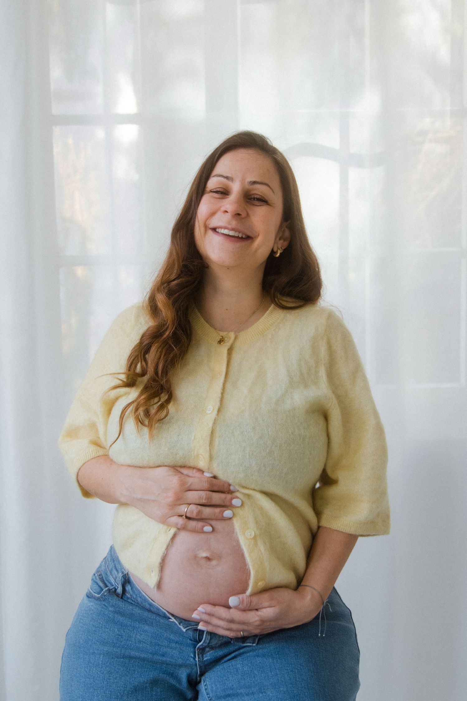Person in yellow top and jeans poses joyfully during maternity photoshoot with white curtain backdrop.