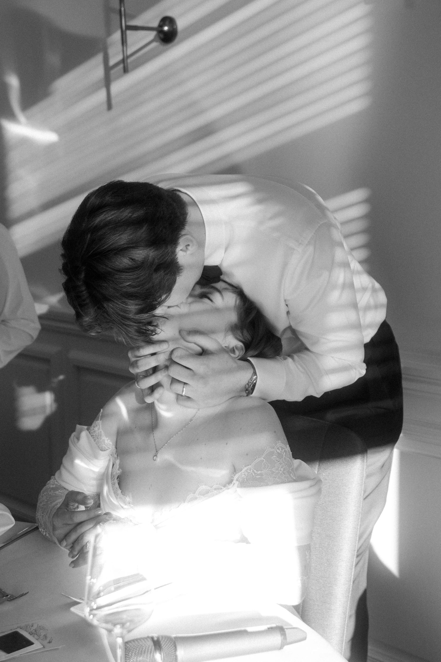 Romantic black and white photo of groom kissing seated bride in soft natural window light.