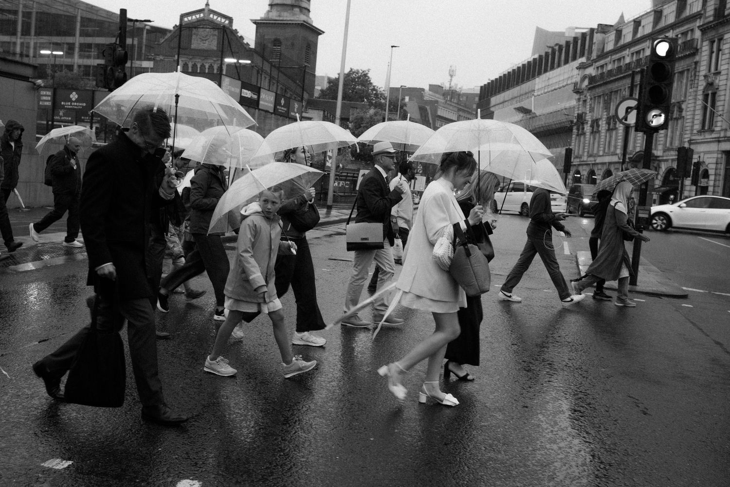 Black and white photo of pedestrians crossing a rainy city street while carrying clear umbrellas.