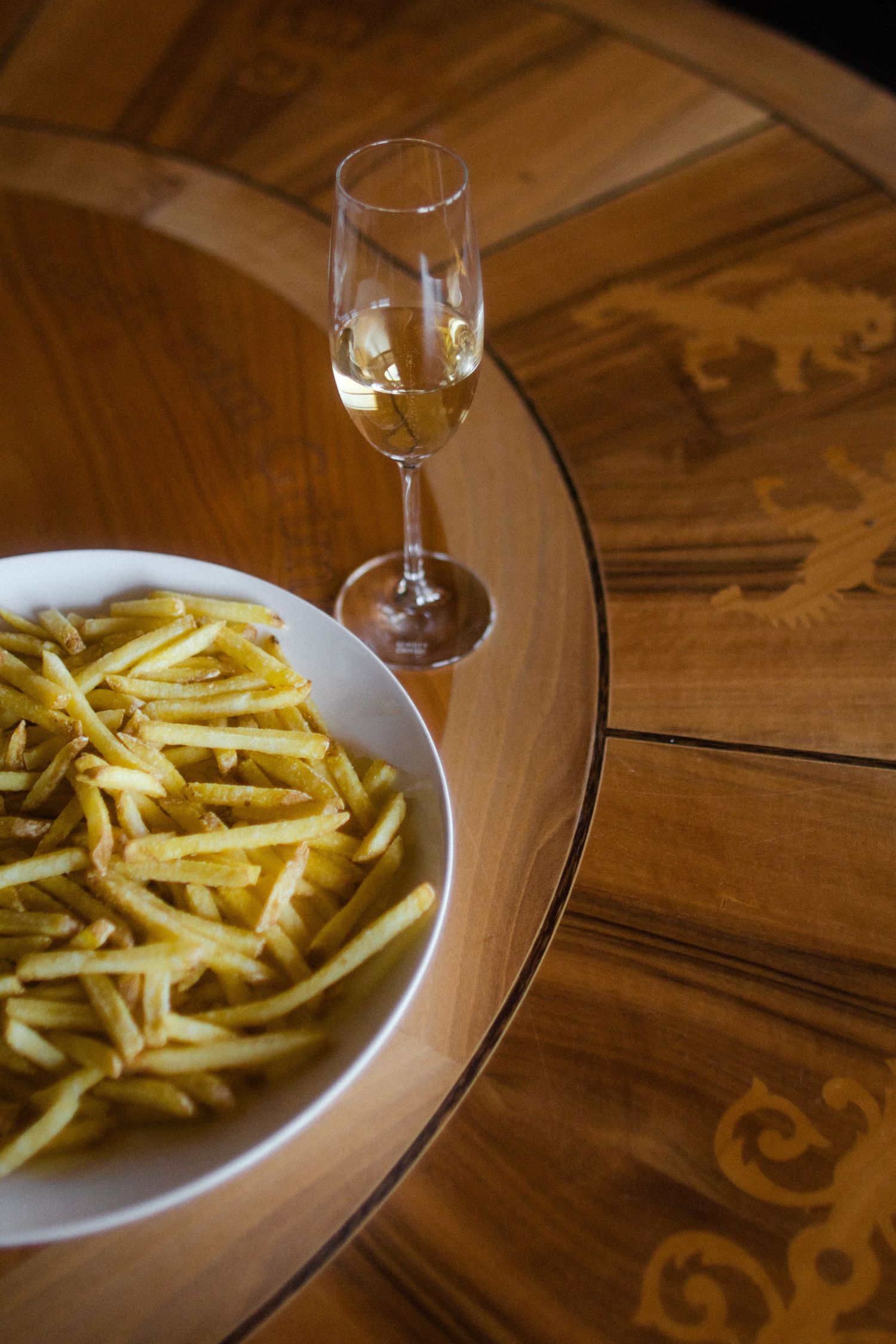 A white bowl of golden french fries beside a champagne flute on an ornate wooden table.