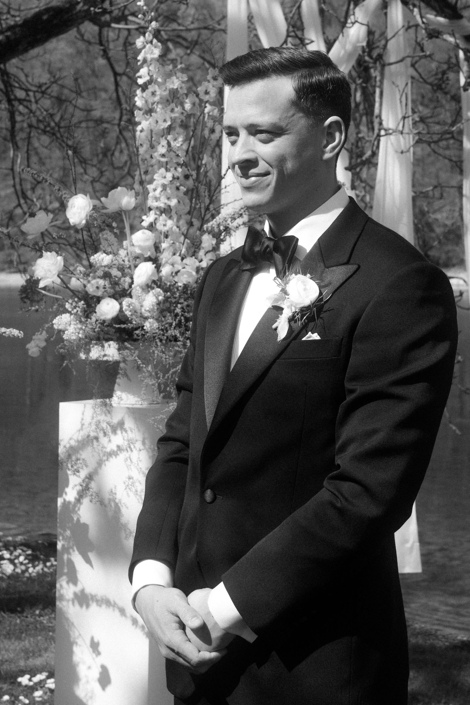 Elegant groom in black tuxedo with boutonniere smiling at outdoor wedding ceremony in black and white.