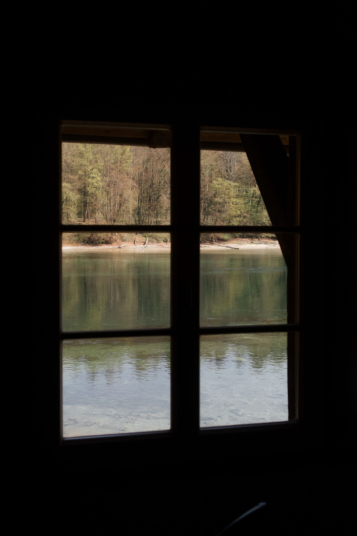 View through a dark window frame overlooking a calm lake with green forest reflections on the water.