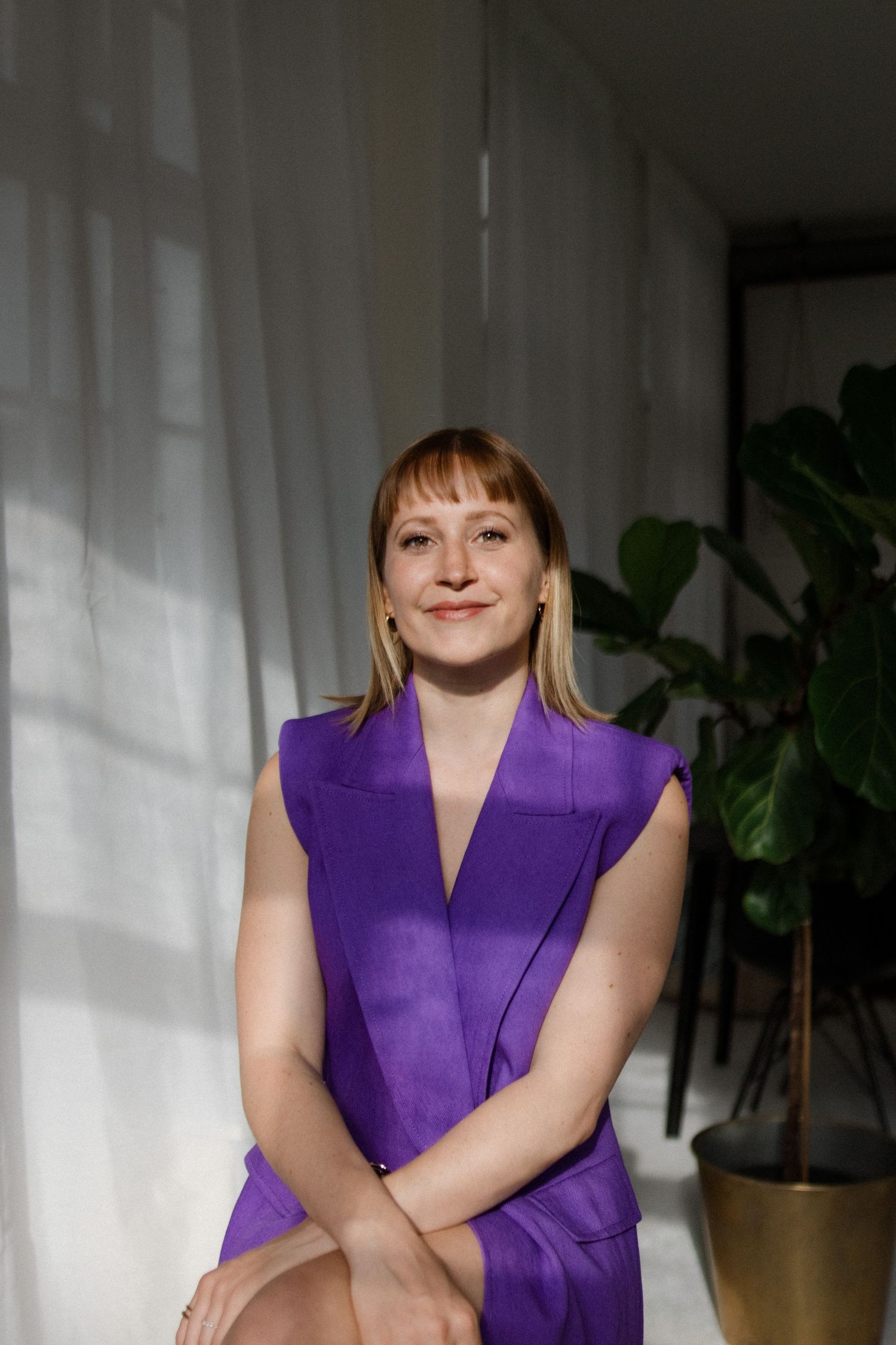 A person wearing a vibrant purple sleeveless dress poses in a bright room with houseplants.
