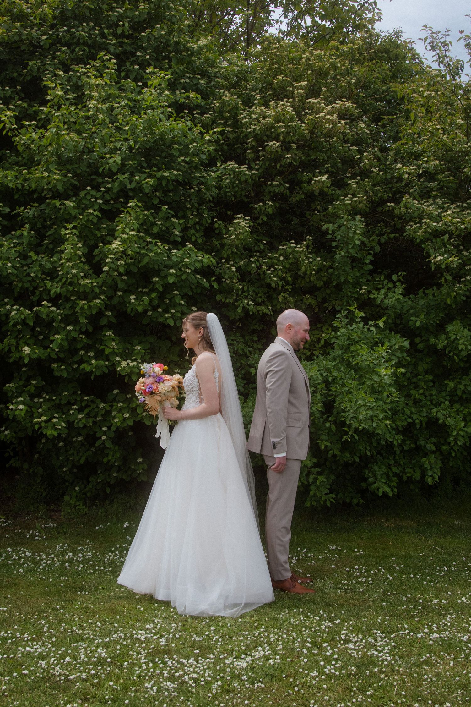 A wedding ceremony unfolds in a garden setting with white flowers scattered across green grass.