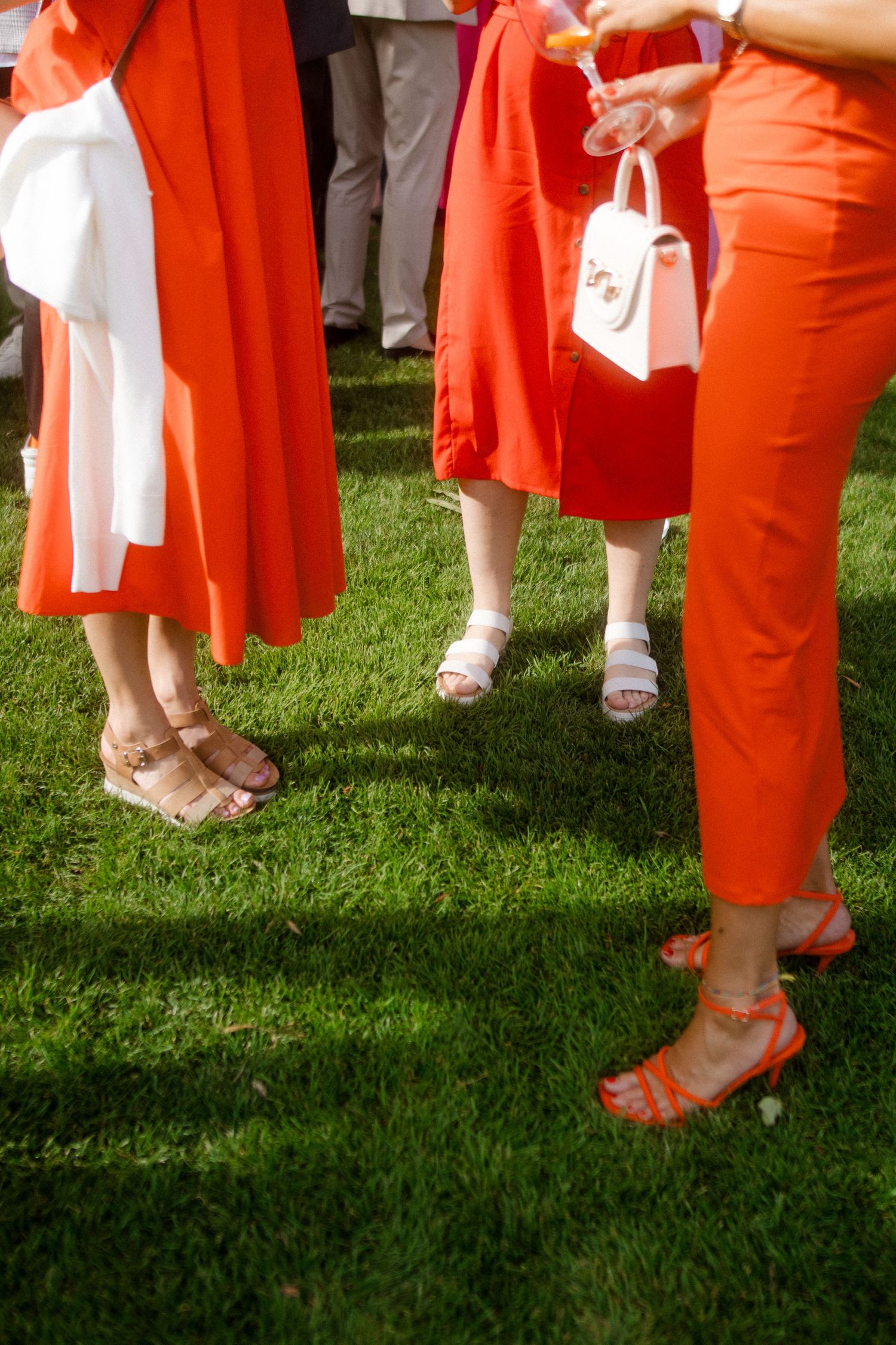 Cropped view of wedding guests in bright coral dresses and sandals standing on grass.