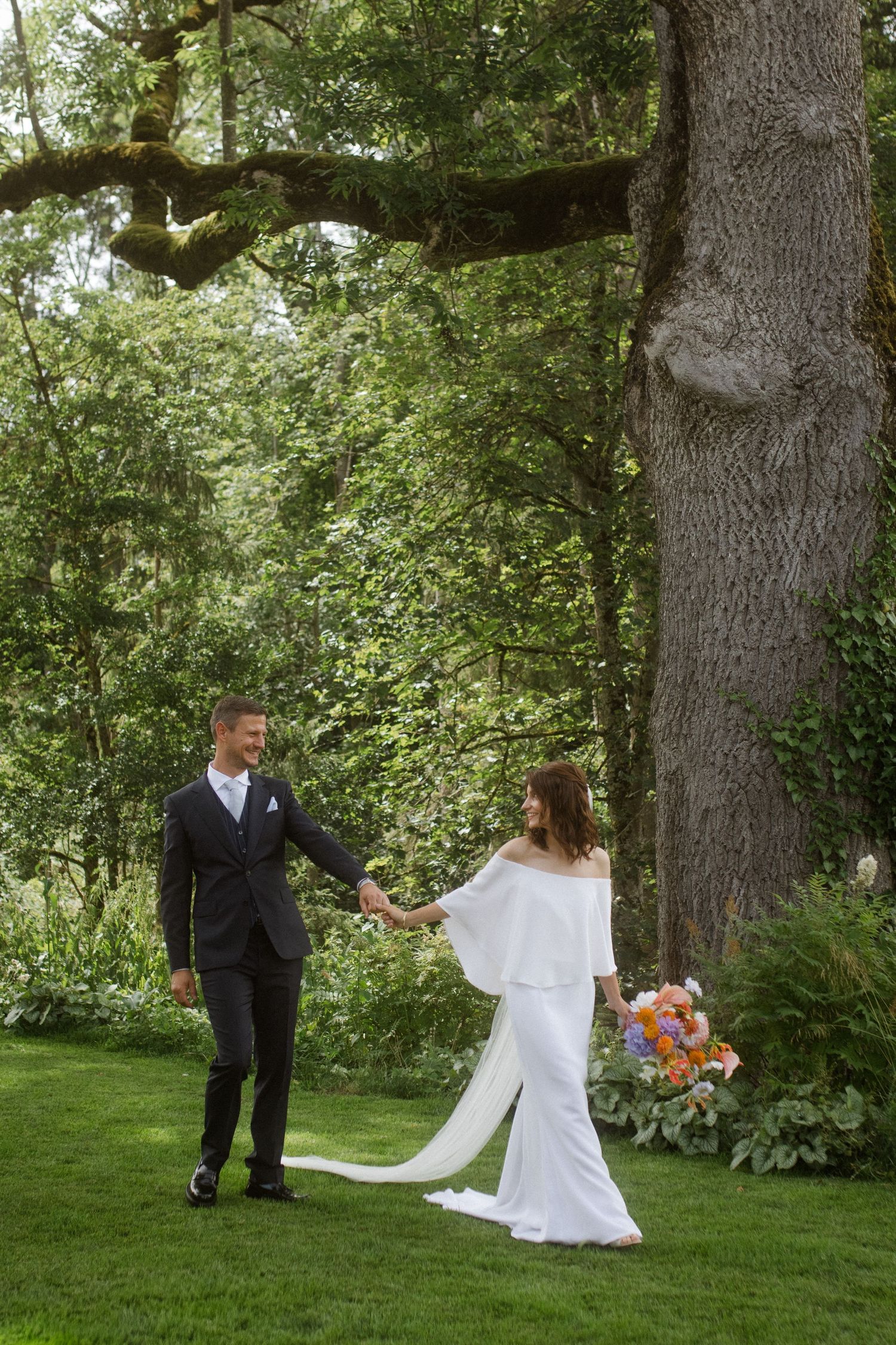 A couple holds hands under a large tree during their outdoor wedding photoshoot.