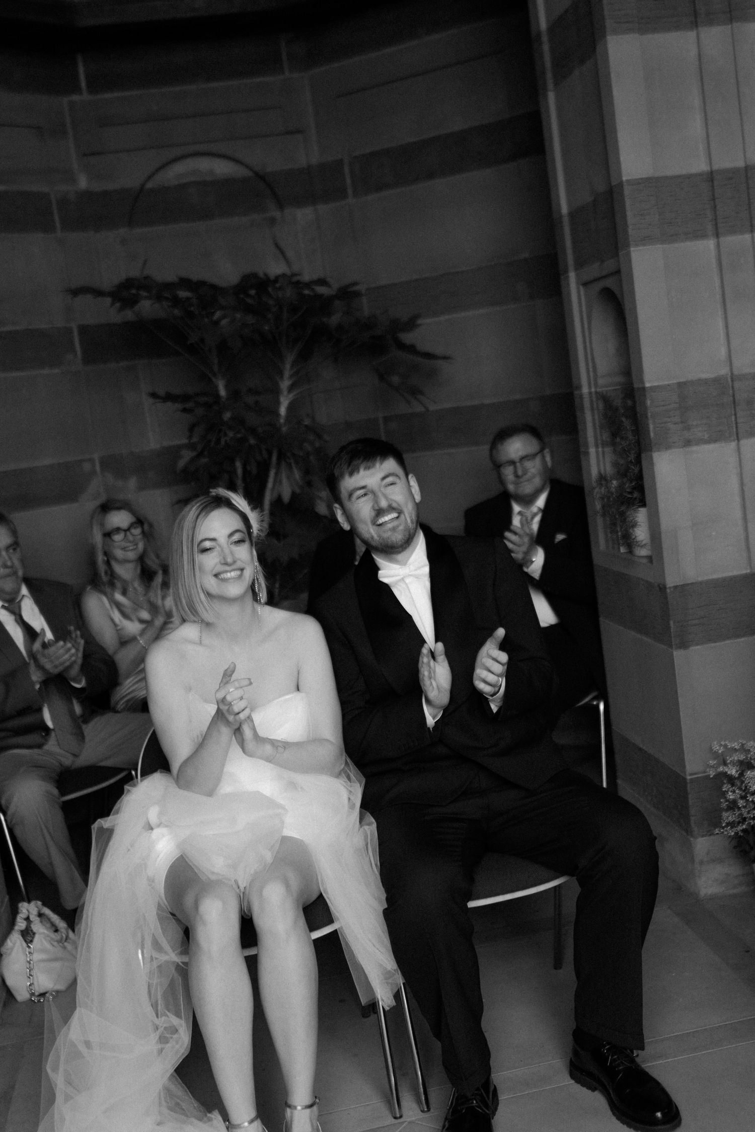 Black and white candid photo of wedding guests sitting in an ornate venue with striped columns.