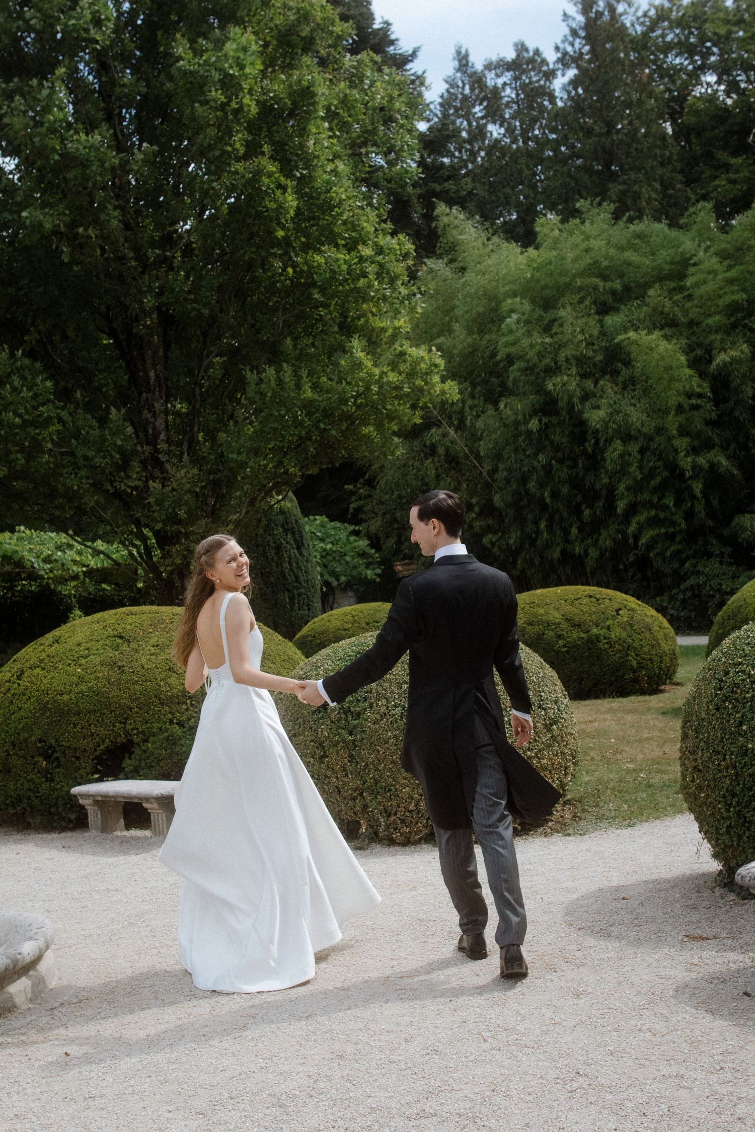 Wedding couple walking along a gravel path surrounded by manicured hedges.