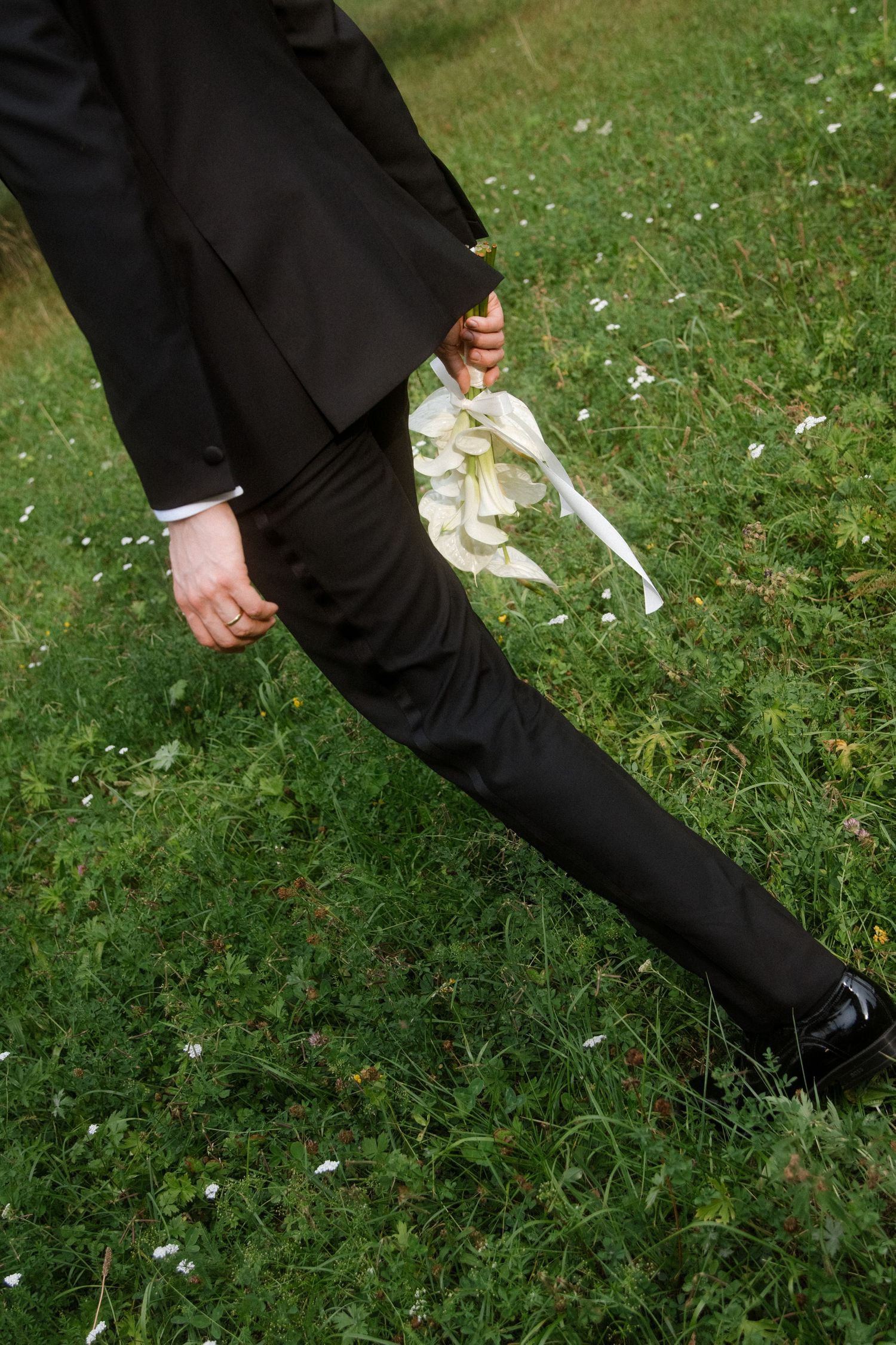 Close-up detail of formal black suit and shoes against green grass with scattered flower petals.