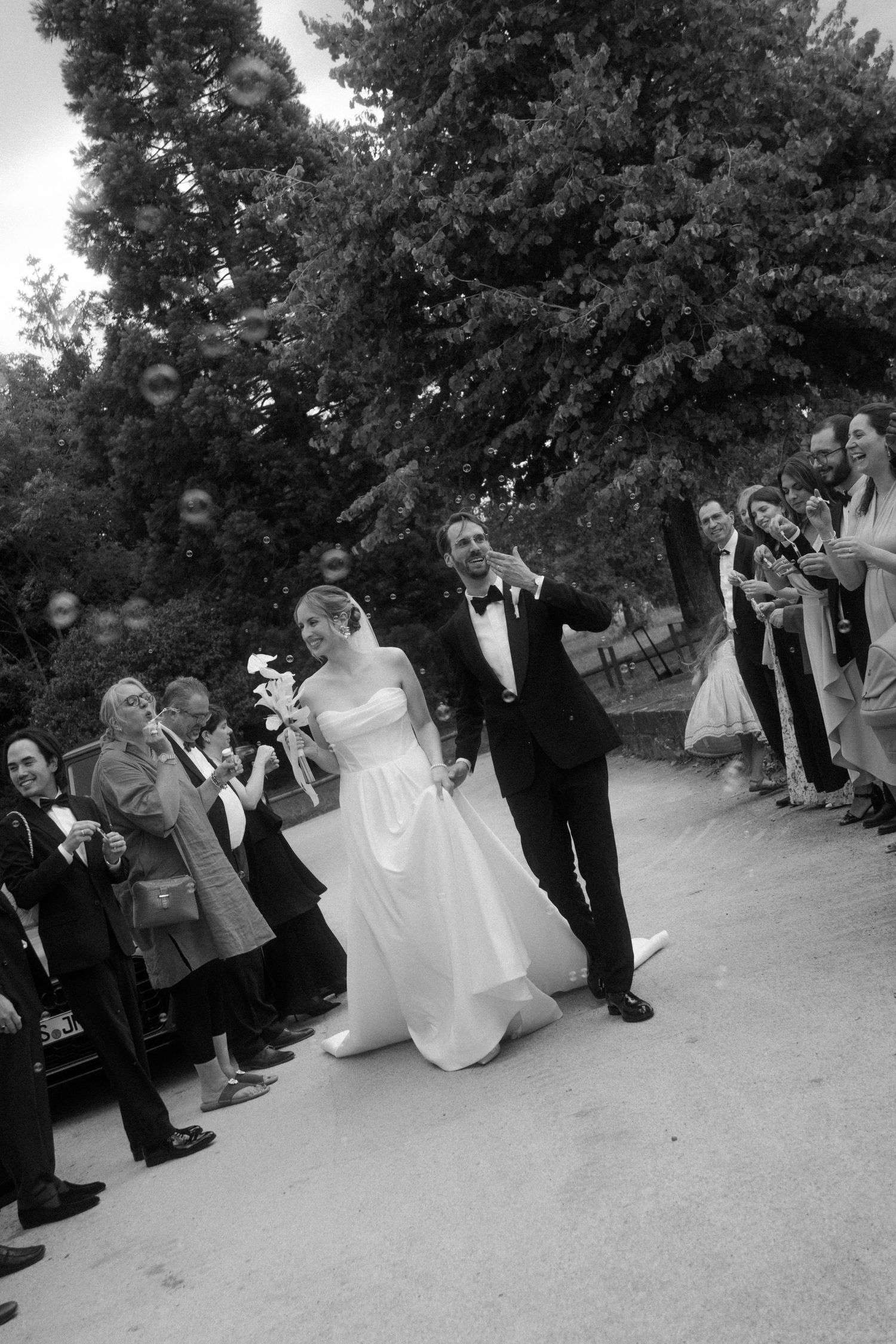 Wedding couple exits while guests line pathway outdoors under leafy trees.