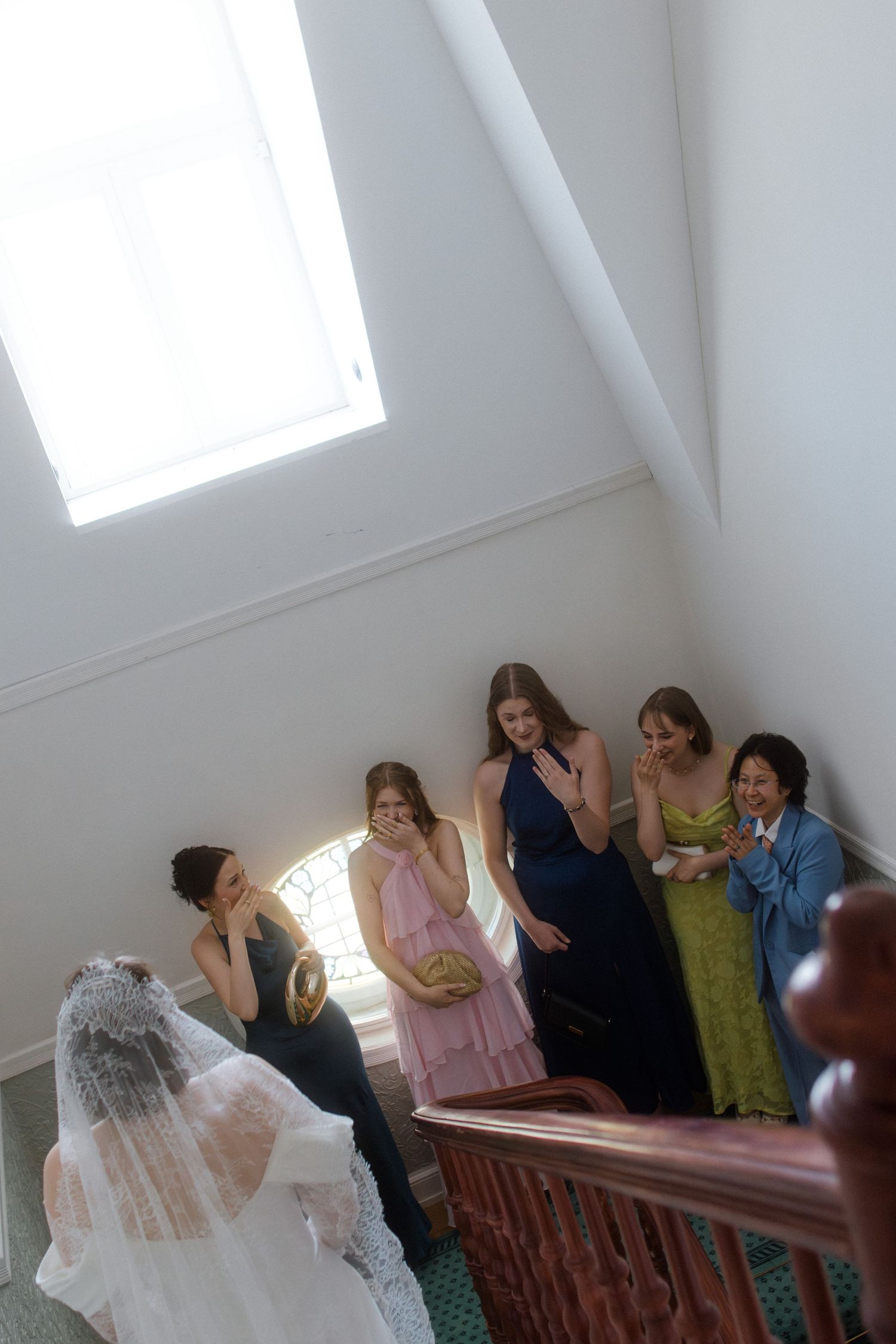 Bride in white gown and veil descends staircase as bridesmaids in colorful dresses react with emotional surprise.