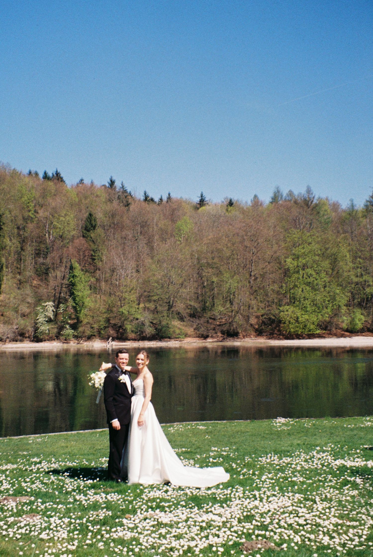 Bride and groom standing on a daisy-covered lawn beside a calm river with lush green forest backdrop on a sunny day.