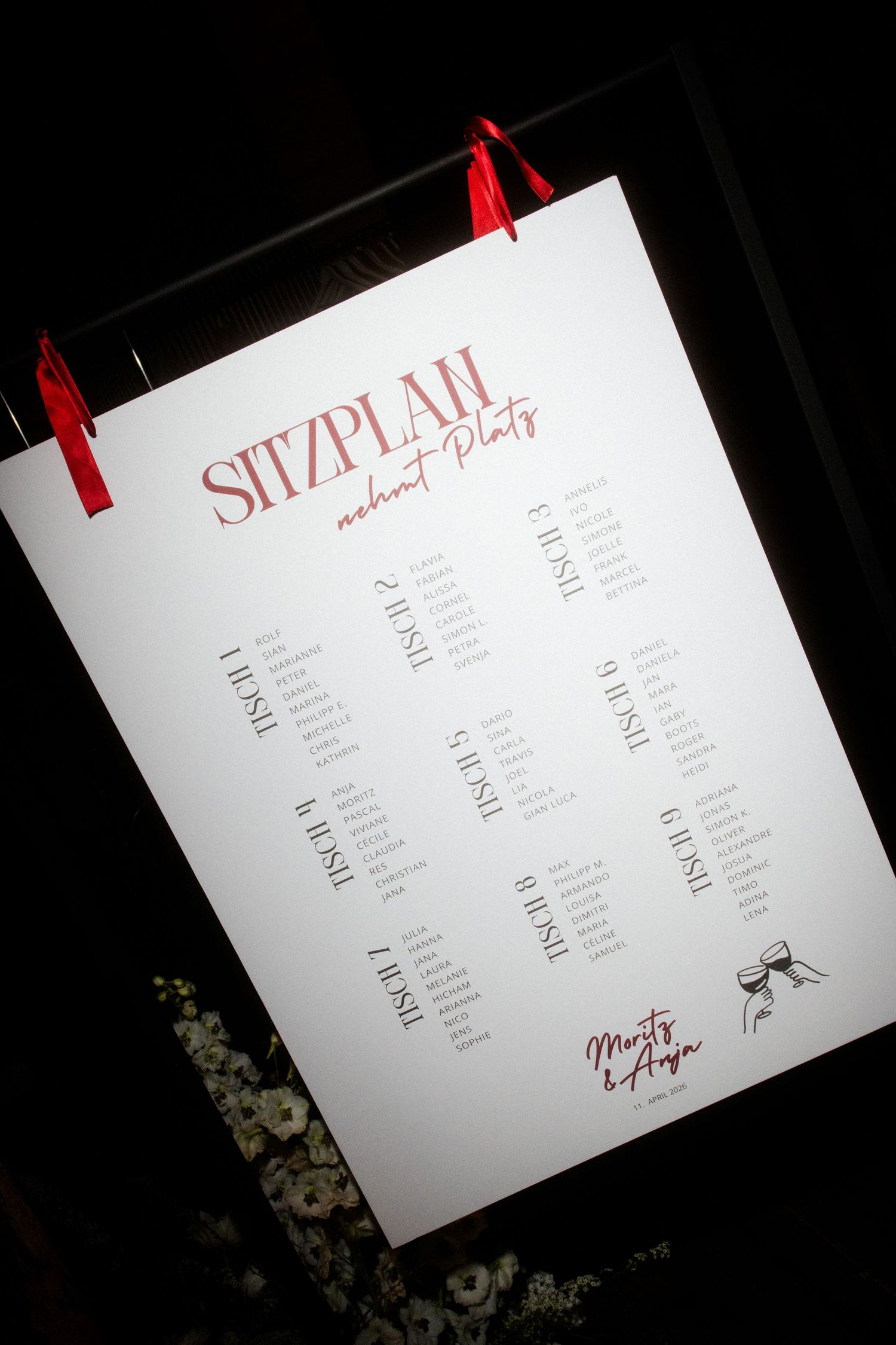 Wedding seating chart titled Sitzplan displayed on a dark board with red ribbon clips.