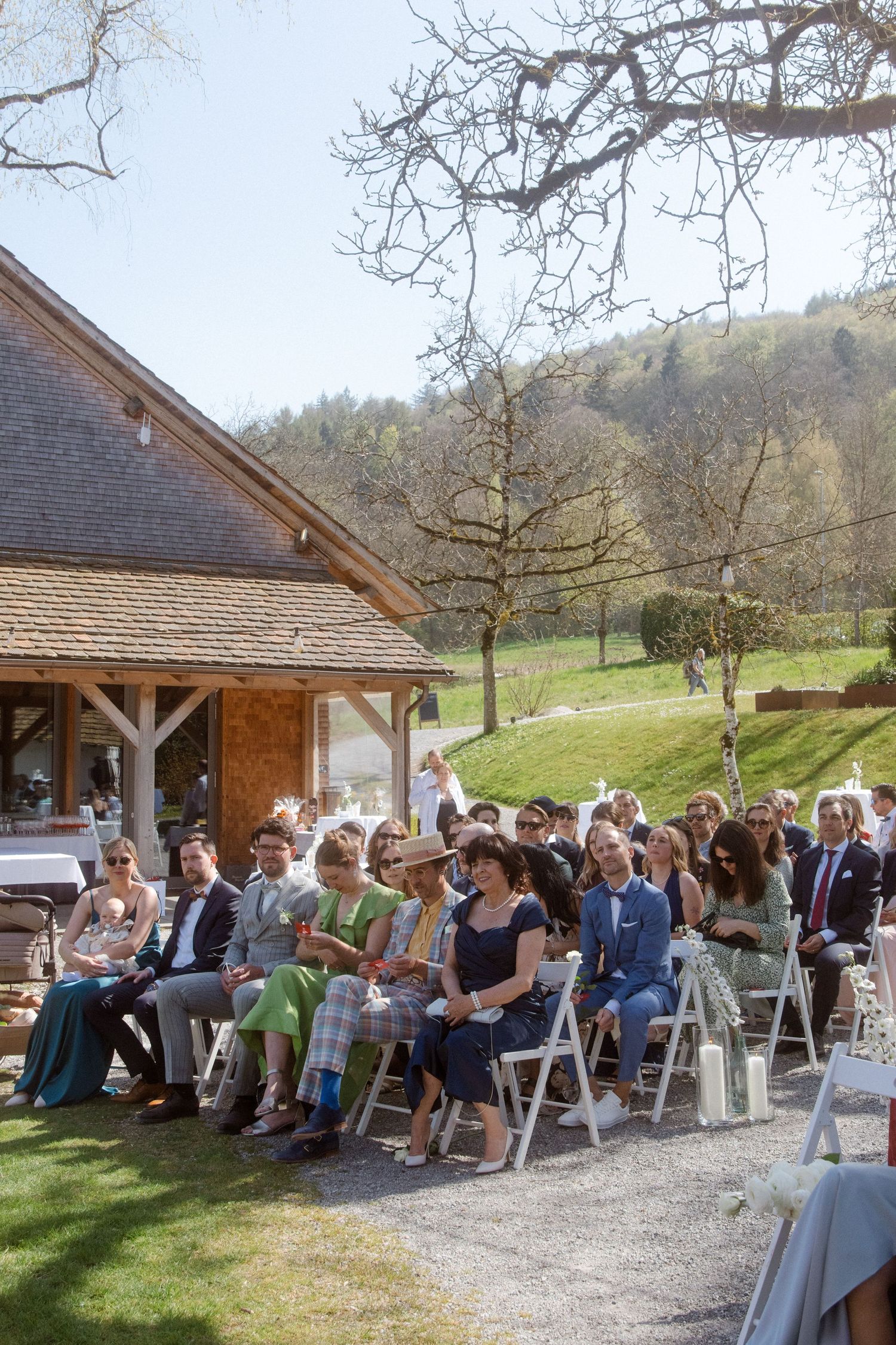 Guests seated on white chairs outdoors at a sunny wedding ceremony beside a wooden barn with rolling green hills.