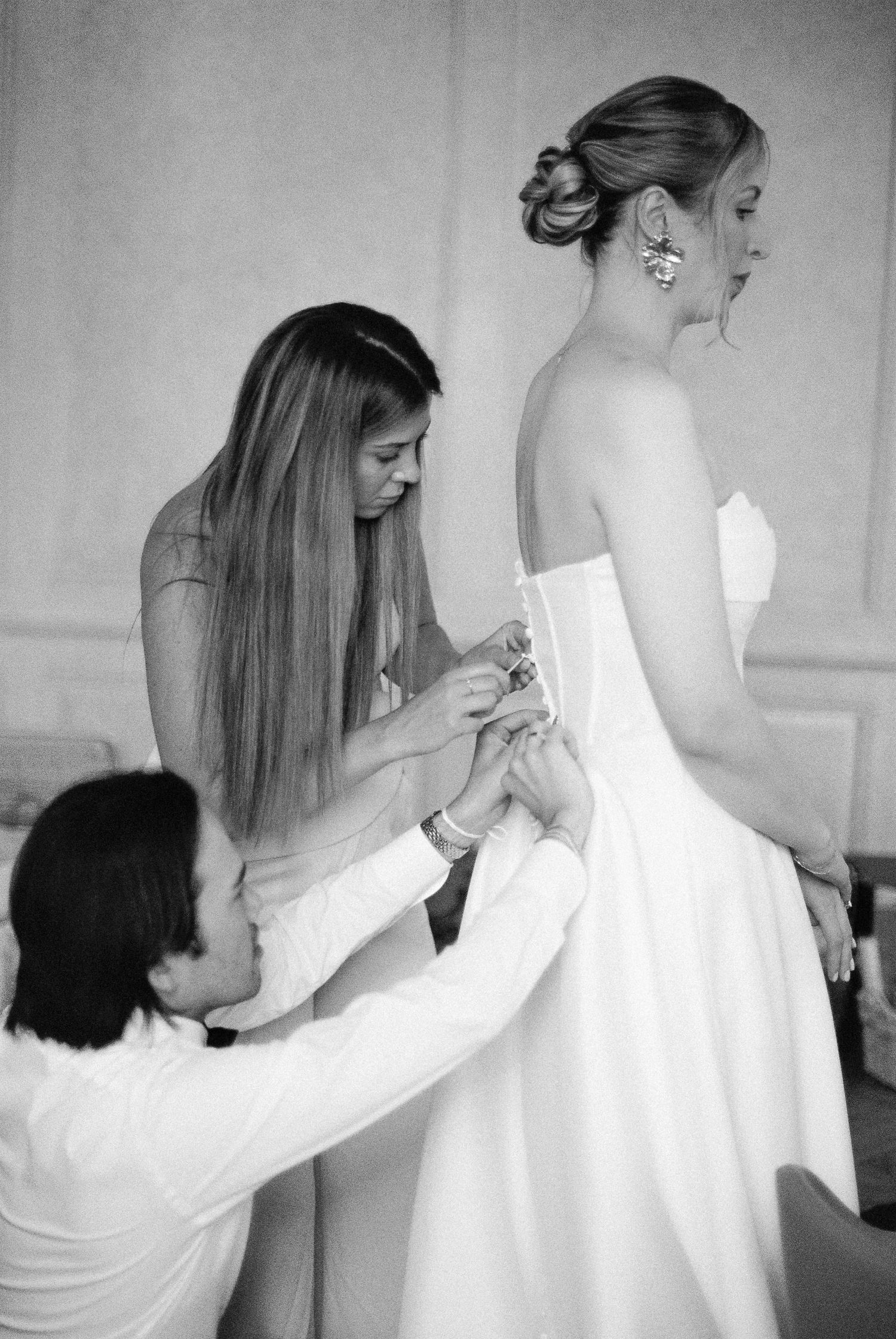 Black and white photo of bride being helped into wedding dress by two bridesmaids before ceremony.