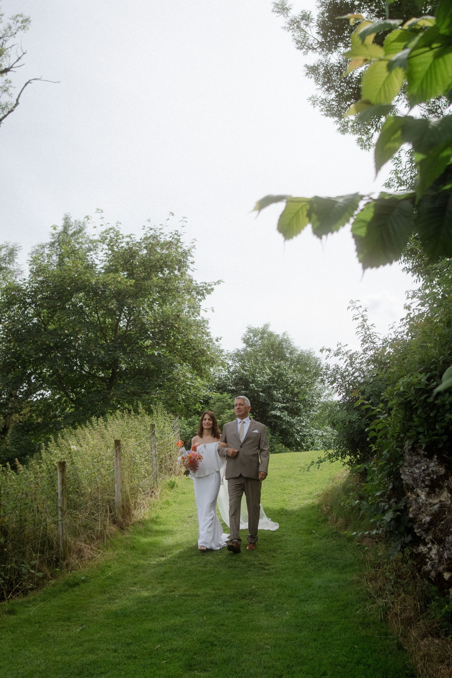 Couple walking along a green garden path surrounded by trees and stone walls.