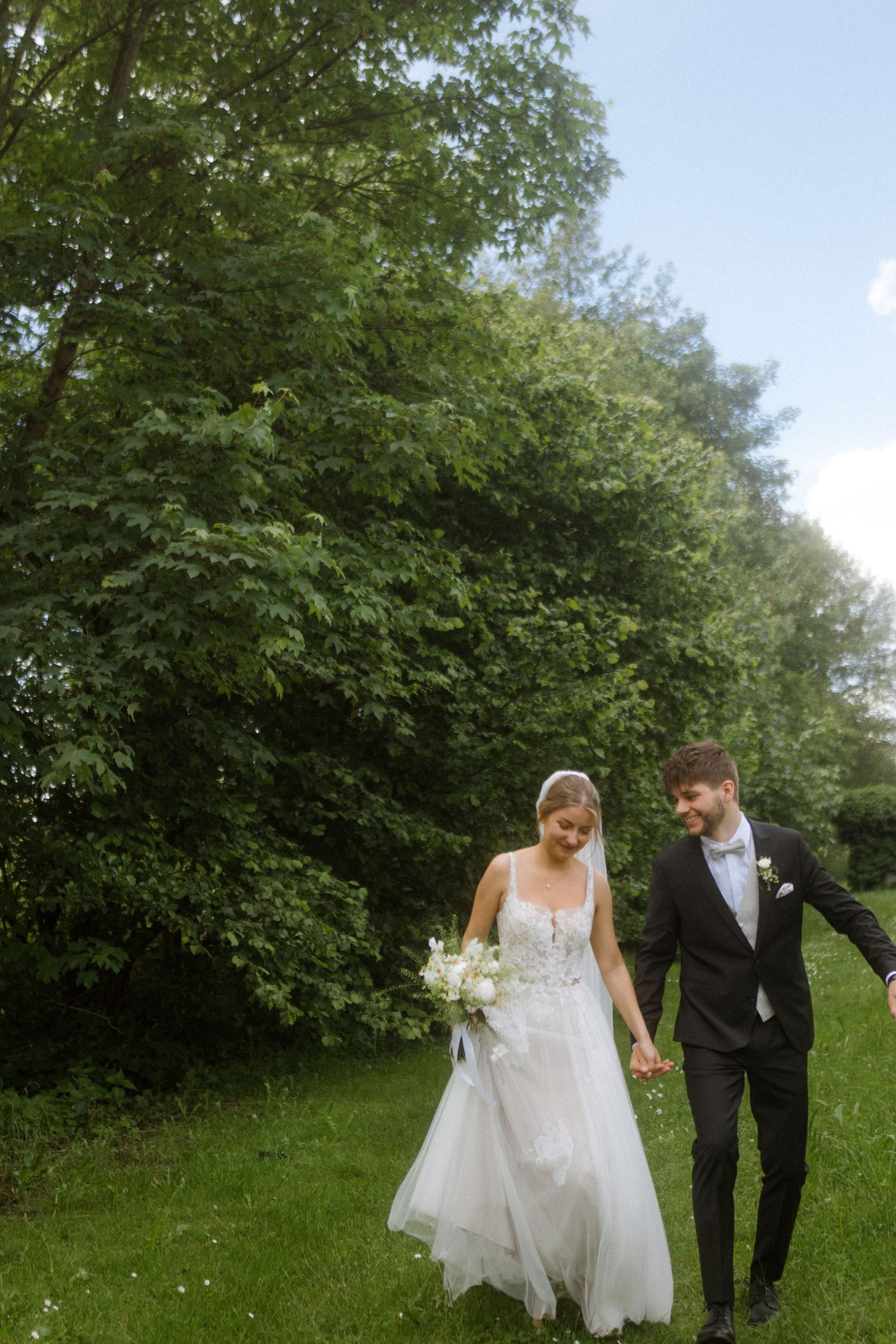 Wedding couple walks hand in hand through a lush green garden path on a sunny summer day.