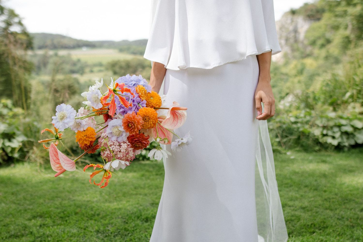 Close-up detail of a white wedding dress and vibrant orange and pink floral bouquet against green landscape.