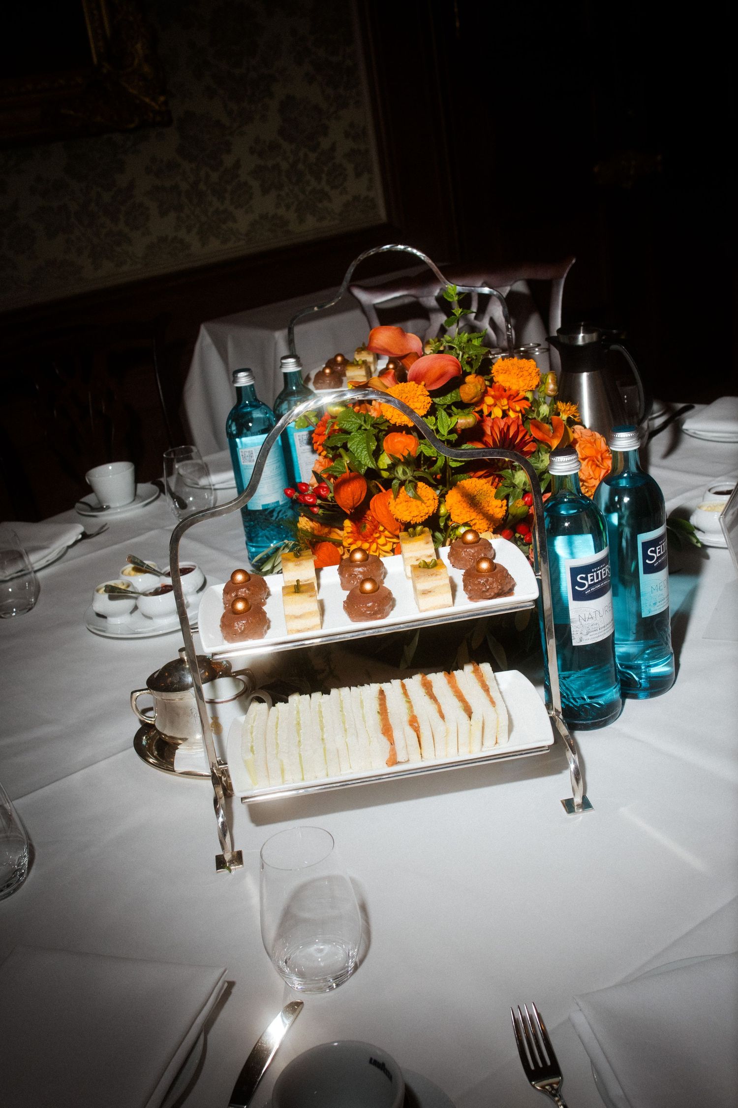 Elegant tiered serving platter with appetizers, sandwiches and drinks on a formal dining table.