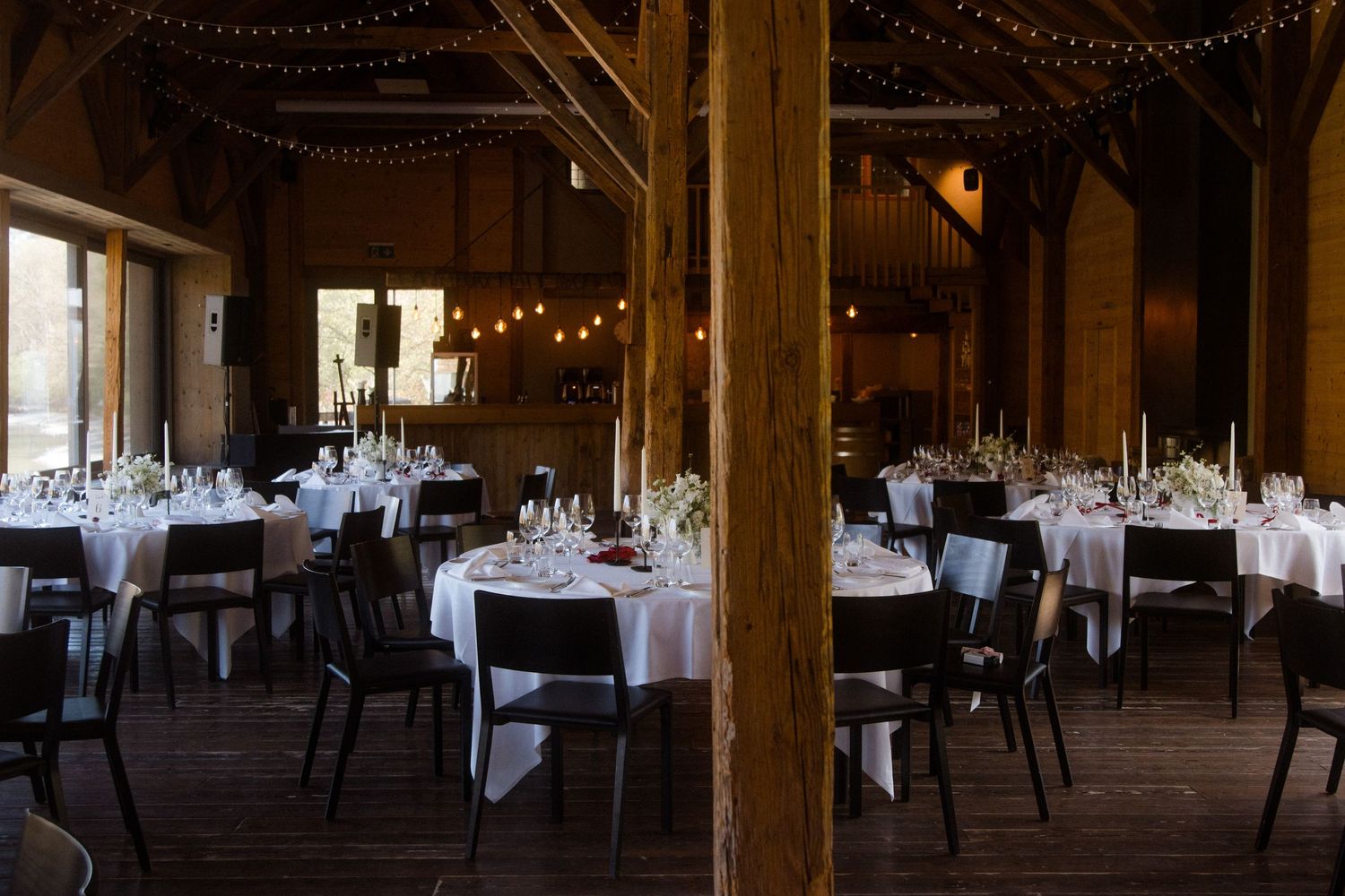 Rustic barn wedding reception hall with round tables, dark chairs, candlelit centerpieces, and warm string lights overhead.
