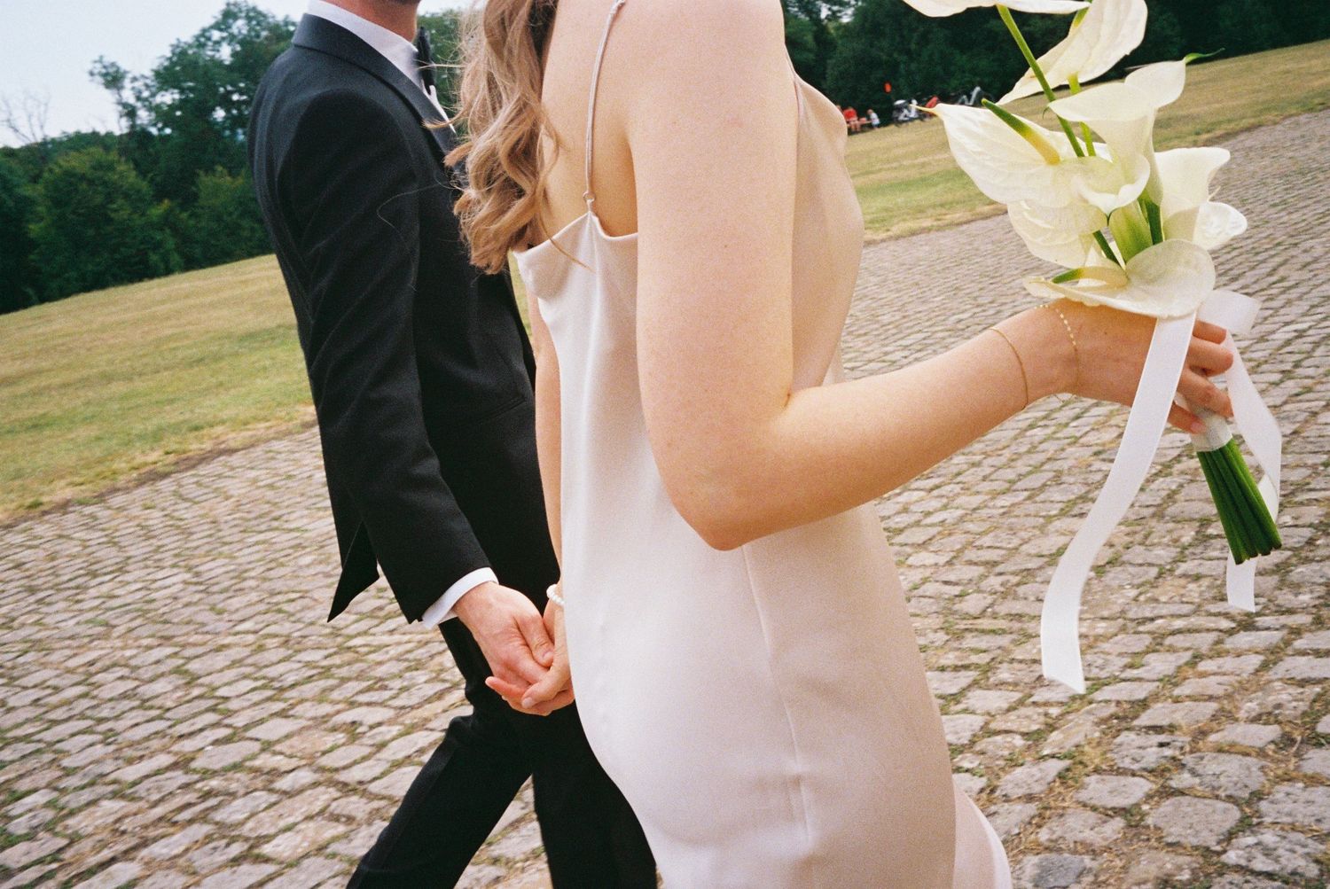 A couple walks along a cobblestone path with a white lily bouquet during their wedding ceremony.