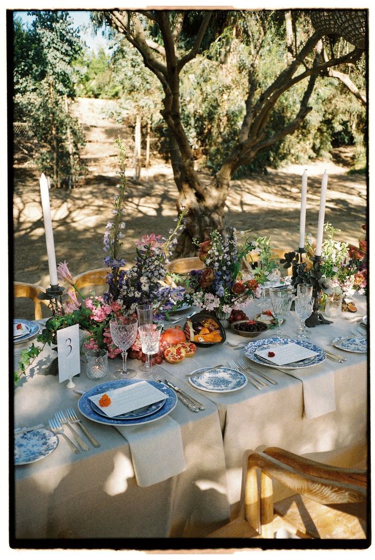 Outdoor wedding table setup under a tree with vintage place settings and floral arrangements.