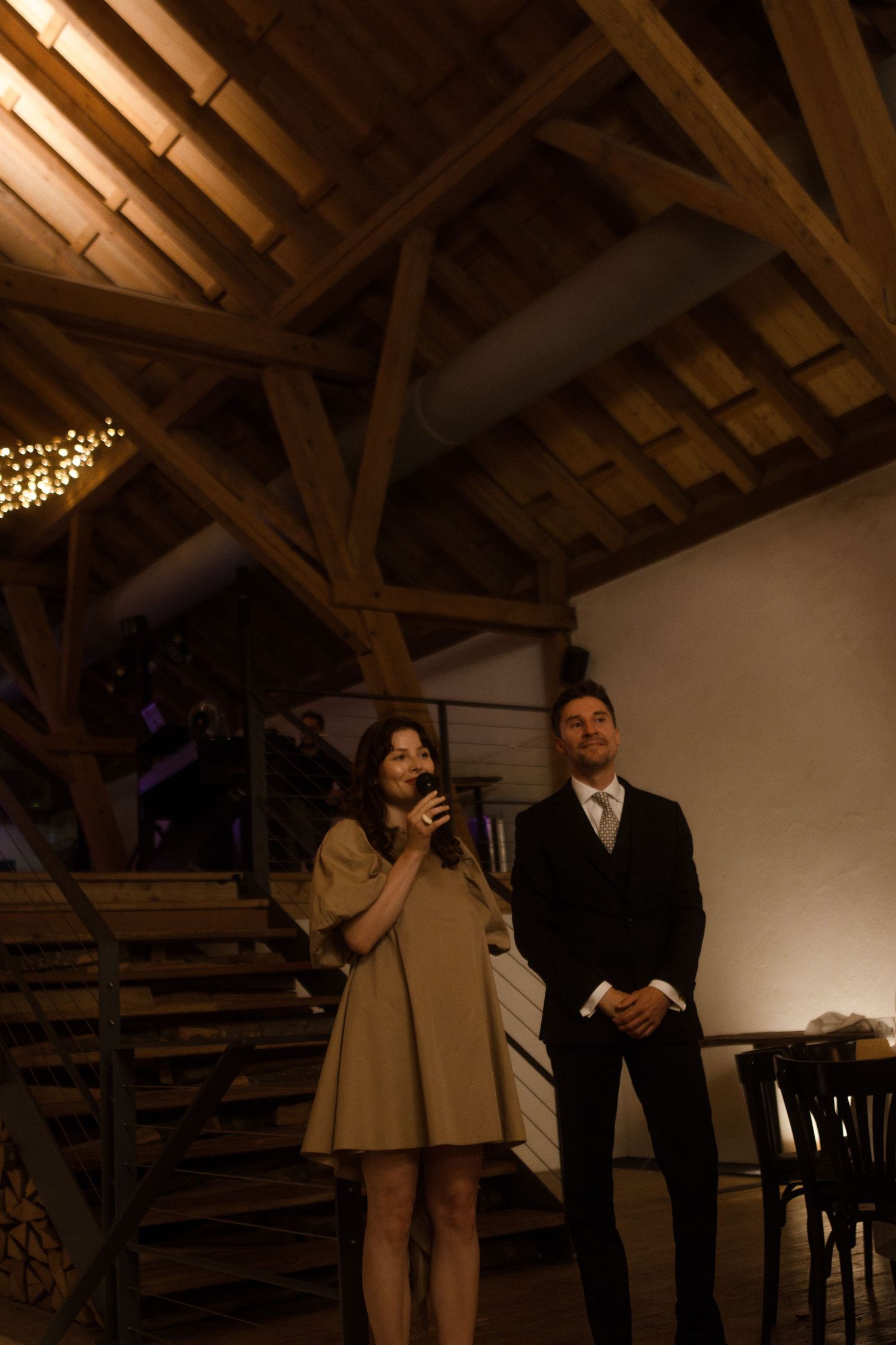 Two people stand on wooden stairs during an indoor event with string lights overhead.