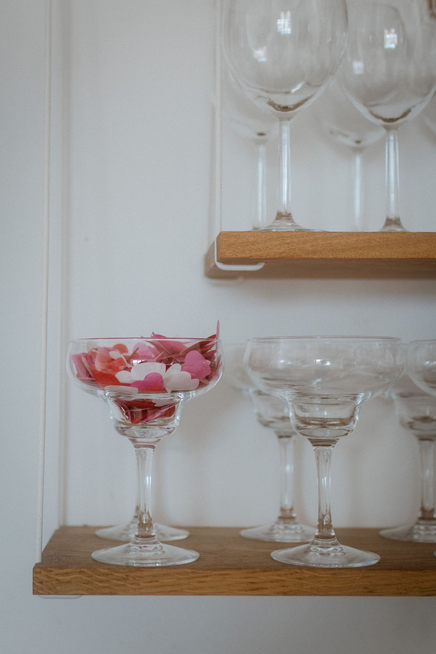 Crystal cocktail glasses displayed on wooden shelves with one containing pink flower petals.