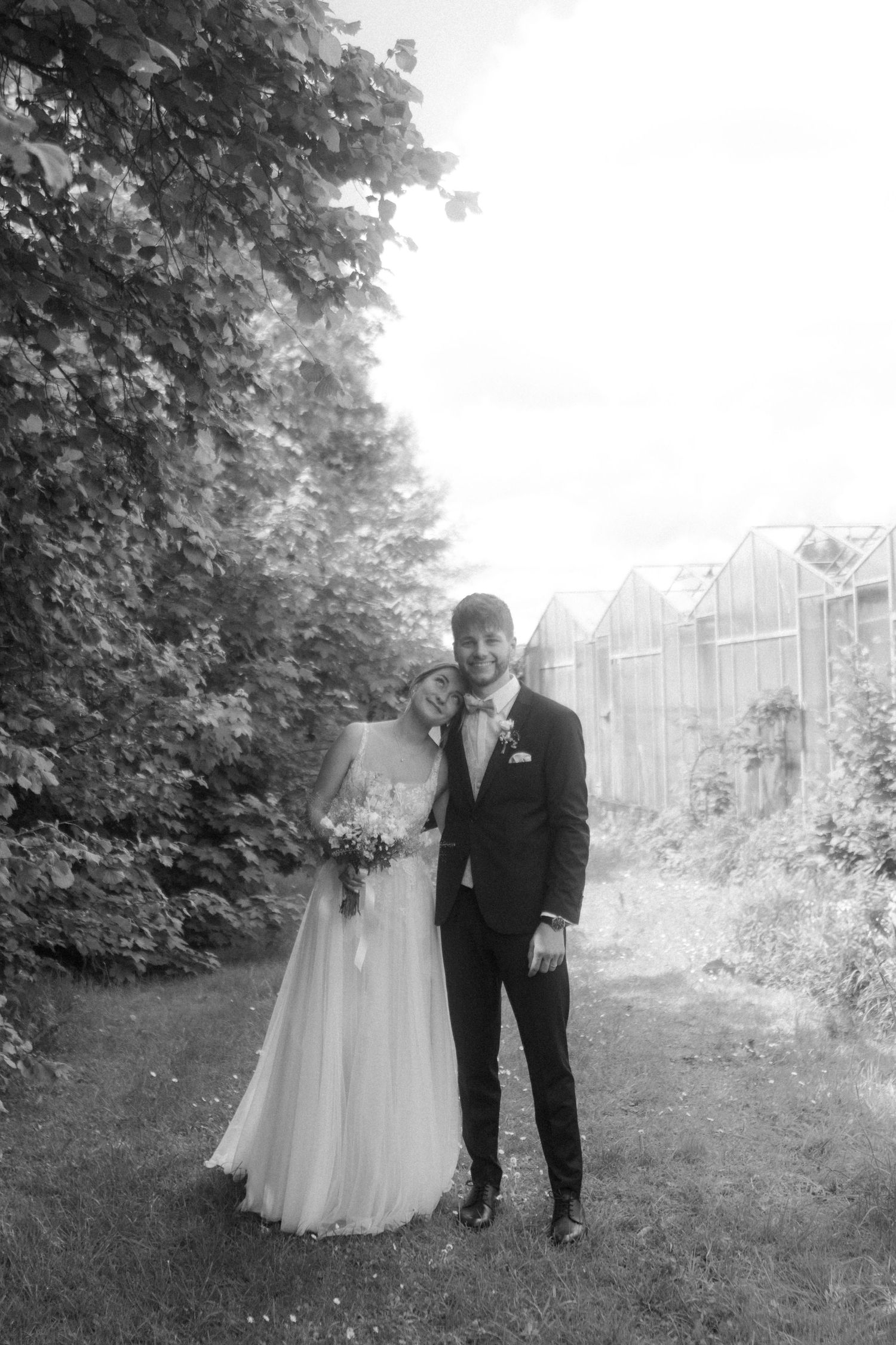 Black and white wedding portrait showing a couple standing on a garden path with greenhouses in the background.