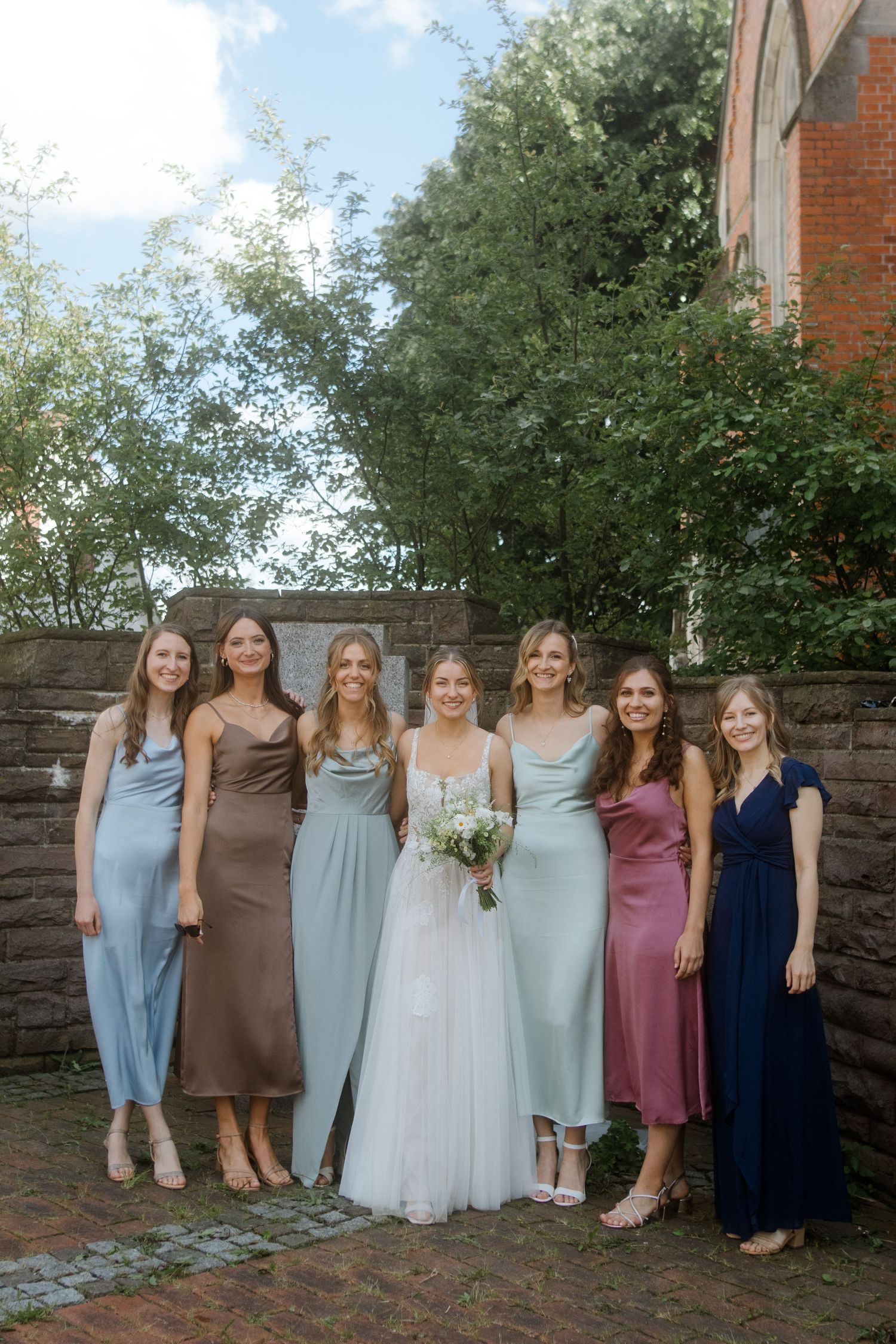 Bridal party in pastel dresses poses against stone wall beside brick church.
