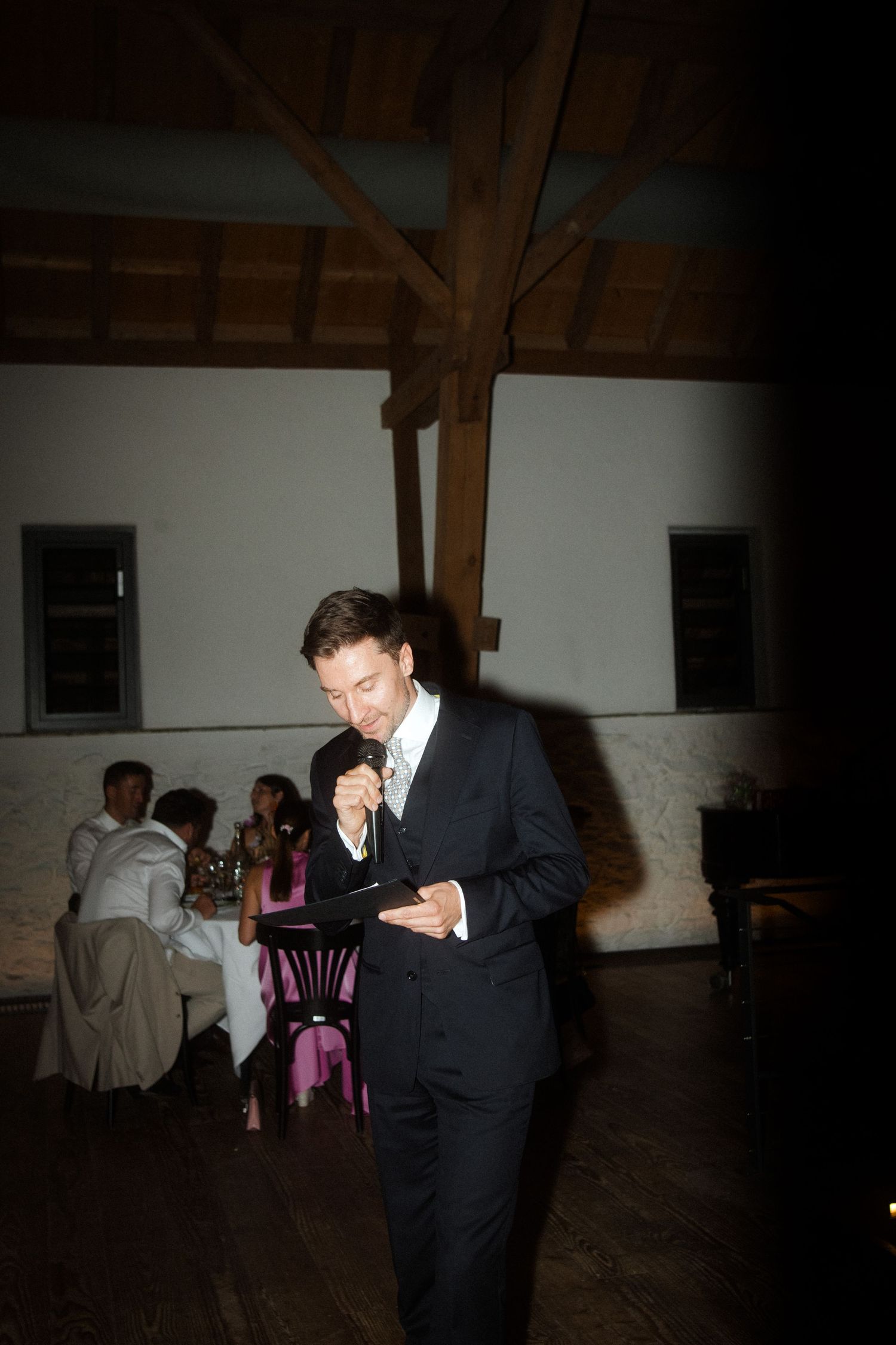 Person in a dark suit standing alone at a reception in a rustic venue.
