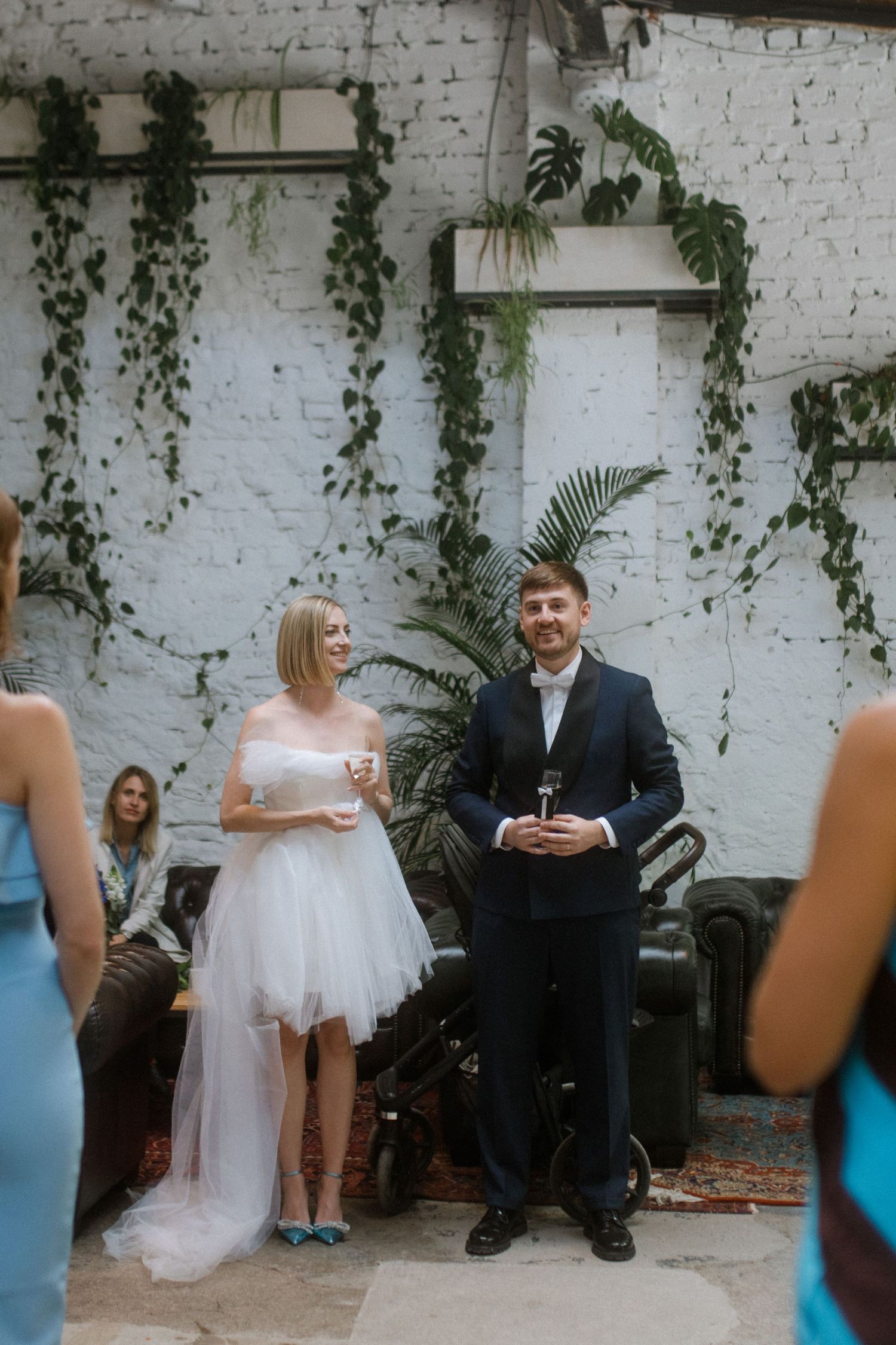A couple gives a toast at their wedding reception in a venue decorated with trailing vines and palm leaves.