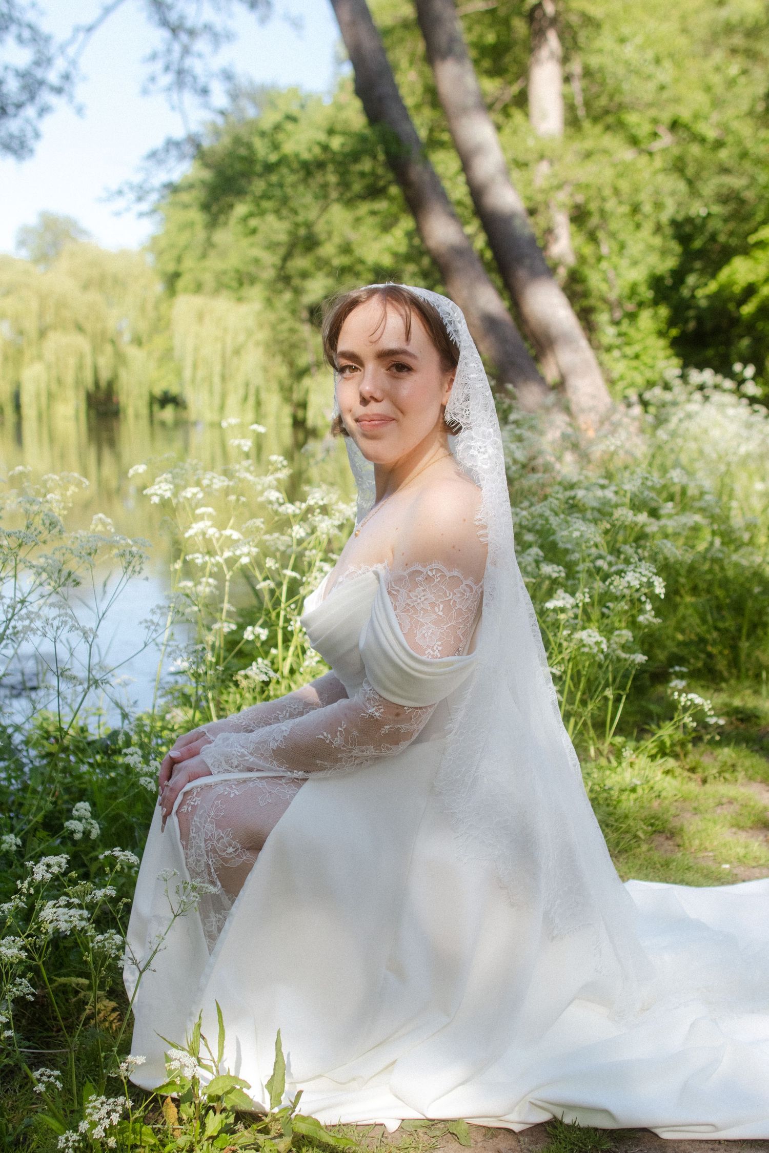 A bride in a white lace gown and veil sits gracefully outdoors near a pond surrounded by lush greenery.