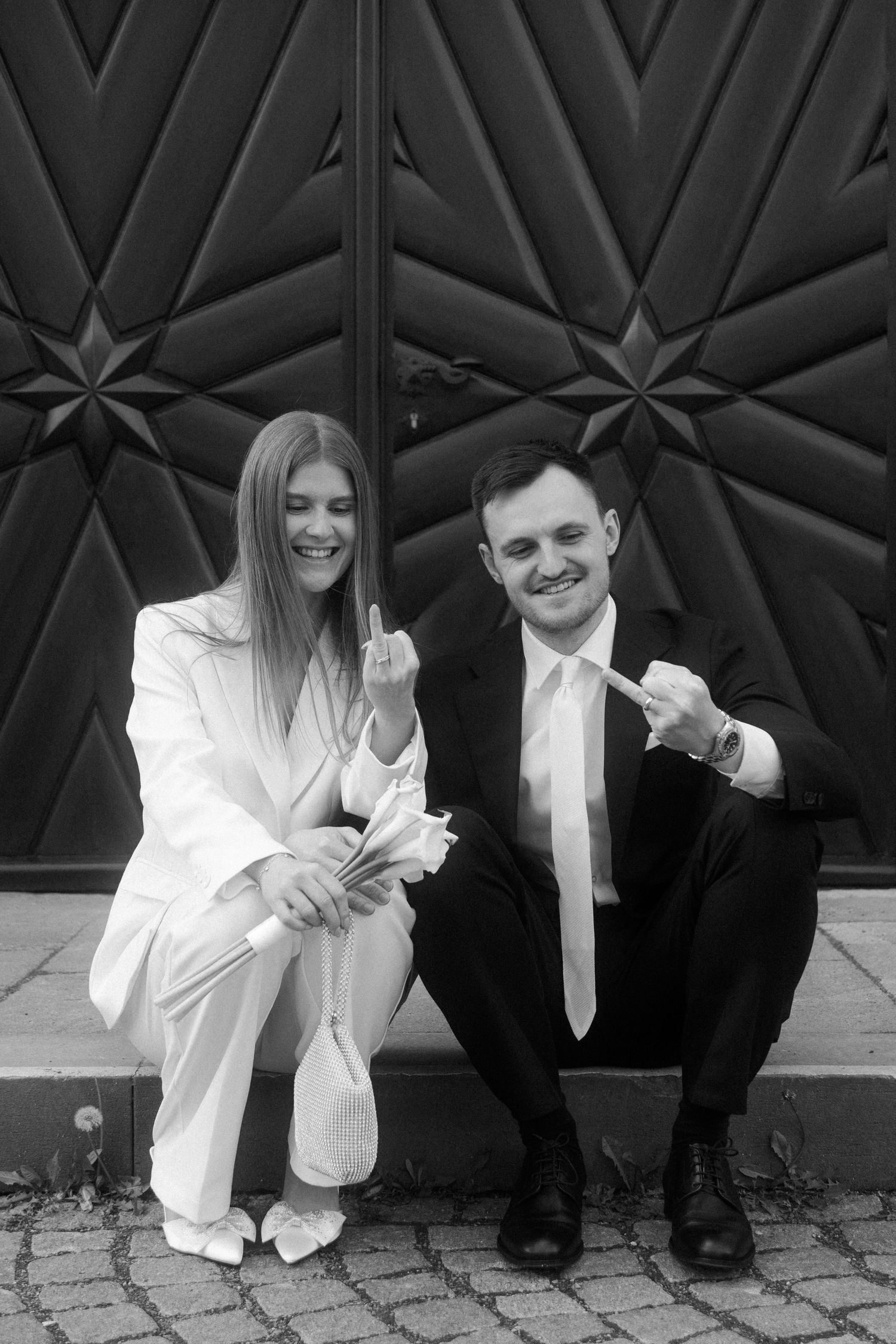 Black and white photo of smiling couple in formal wear sitting outdoors, showing off wedding rings together.