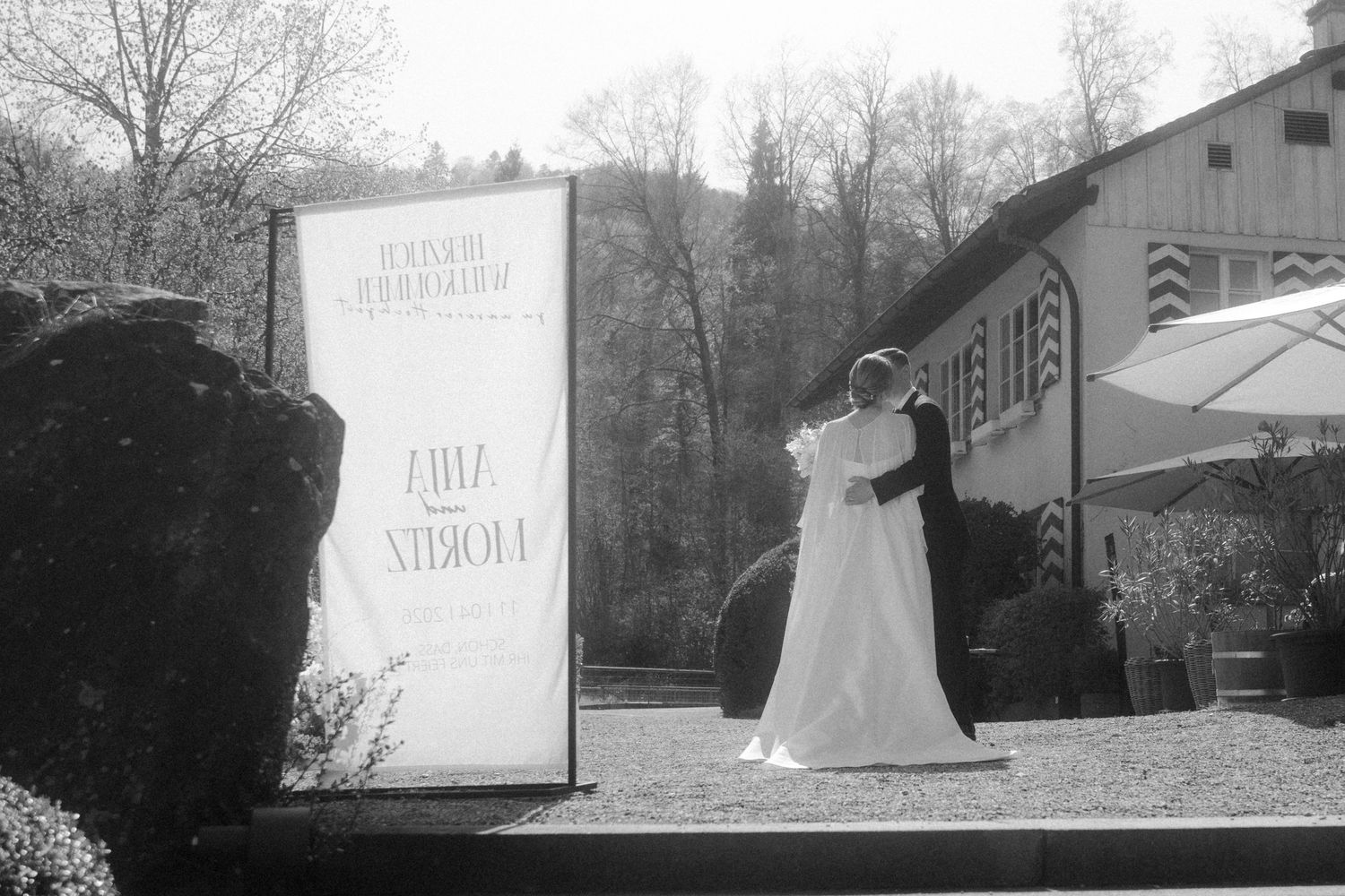 Bride and groom embracing outdoors near a venue sign, surrounded by trees, in a black and white wedding photo.