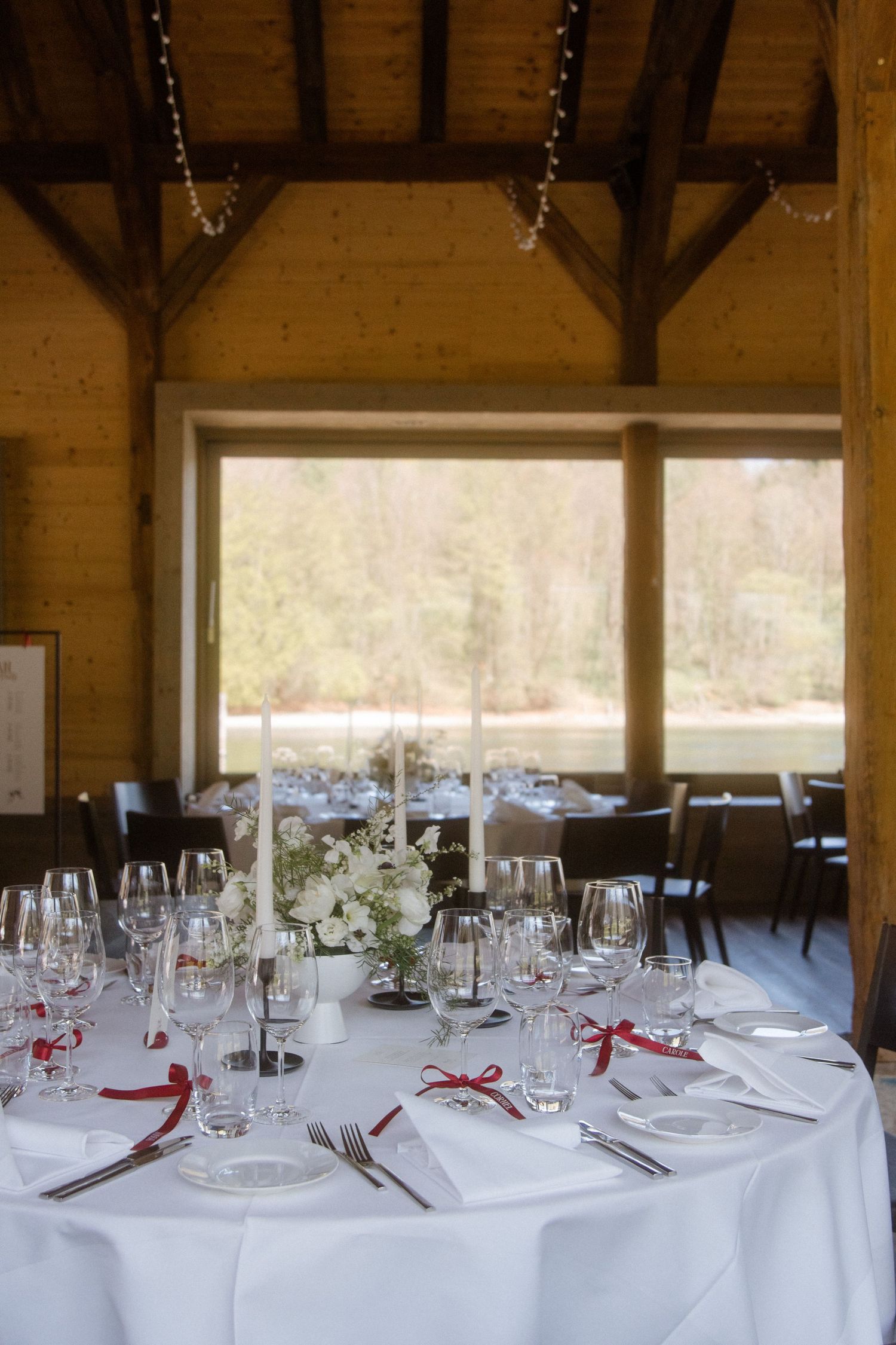 Elegant wedding reception table with white linens, crystal glasses, red ribbon napkins, and floral centerpiece in a rustic wood venue.