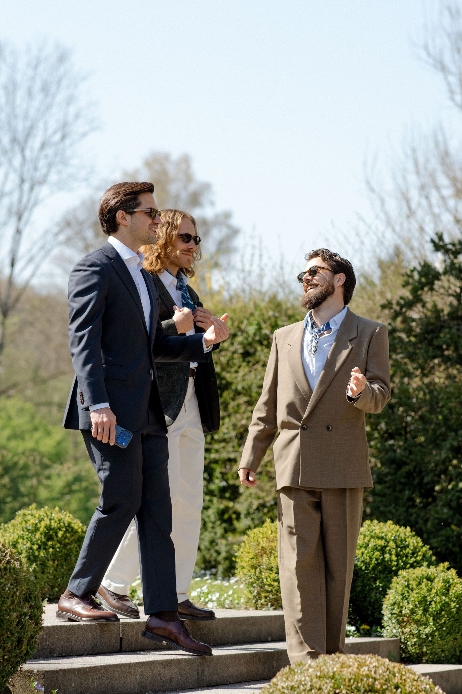 Three well-dressed professionals in suits socializing outdoors on sunny steps surrounded by lush green garden hedges.