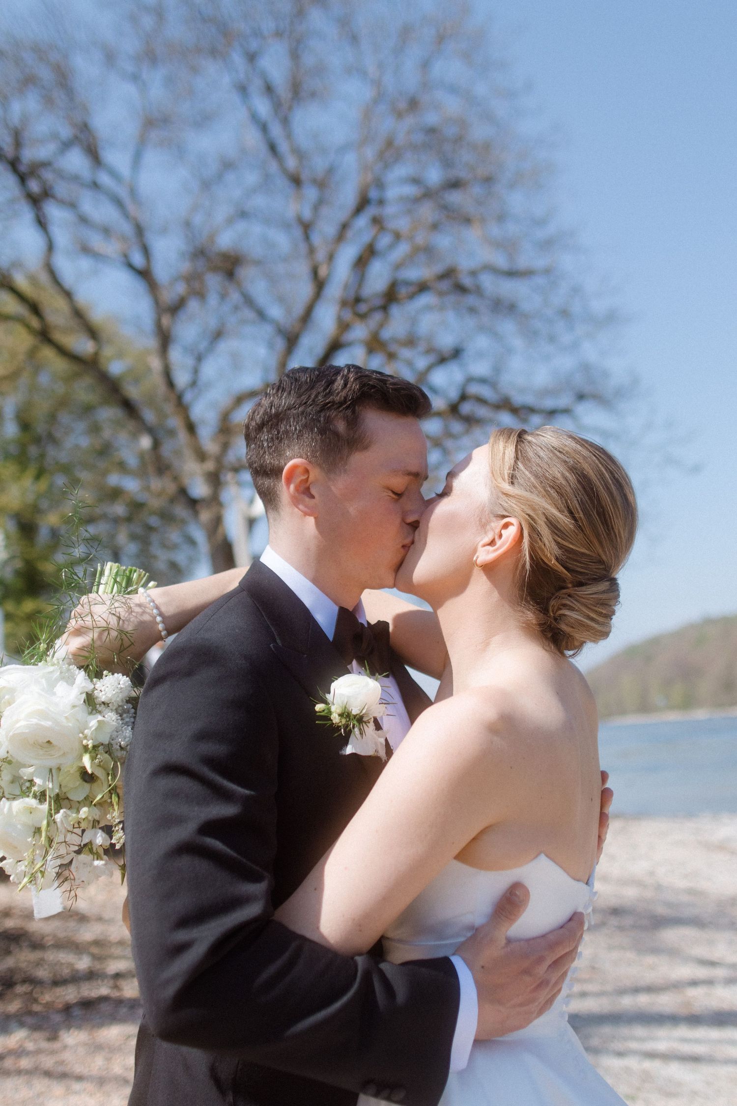 Bride and groom share a romantic kiss outdoors near a lake, with bare trees and blue sky behind them.