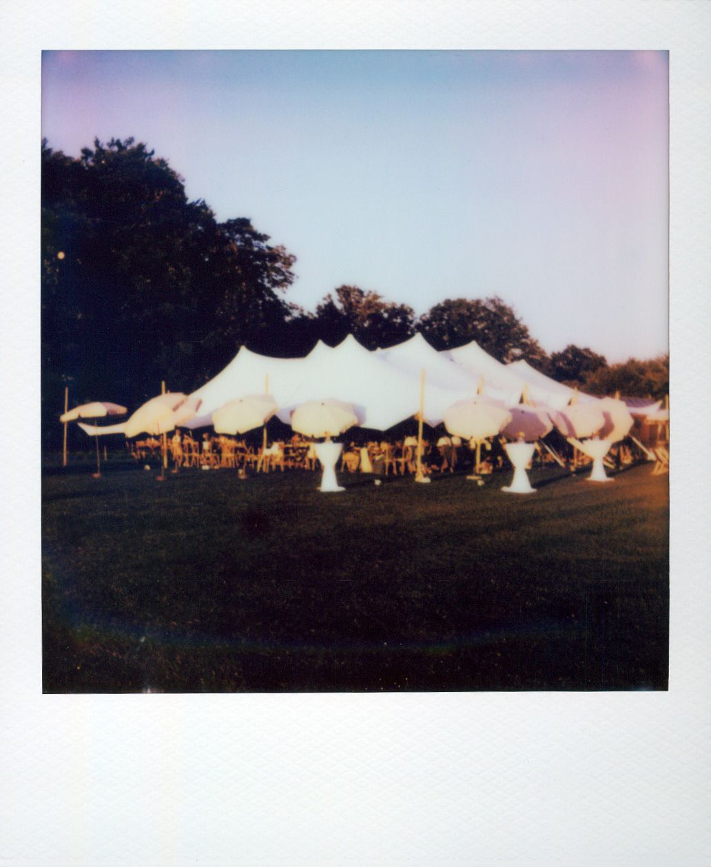 Large white party tents arranged in a line across a grassy field during golden hour with fall foliage visible.