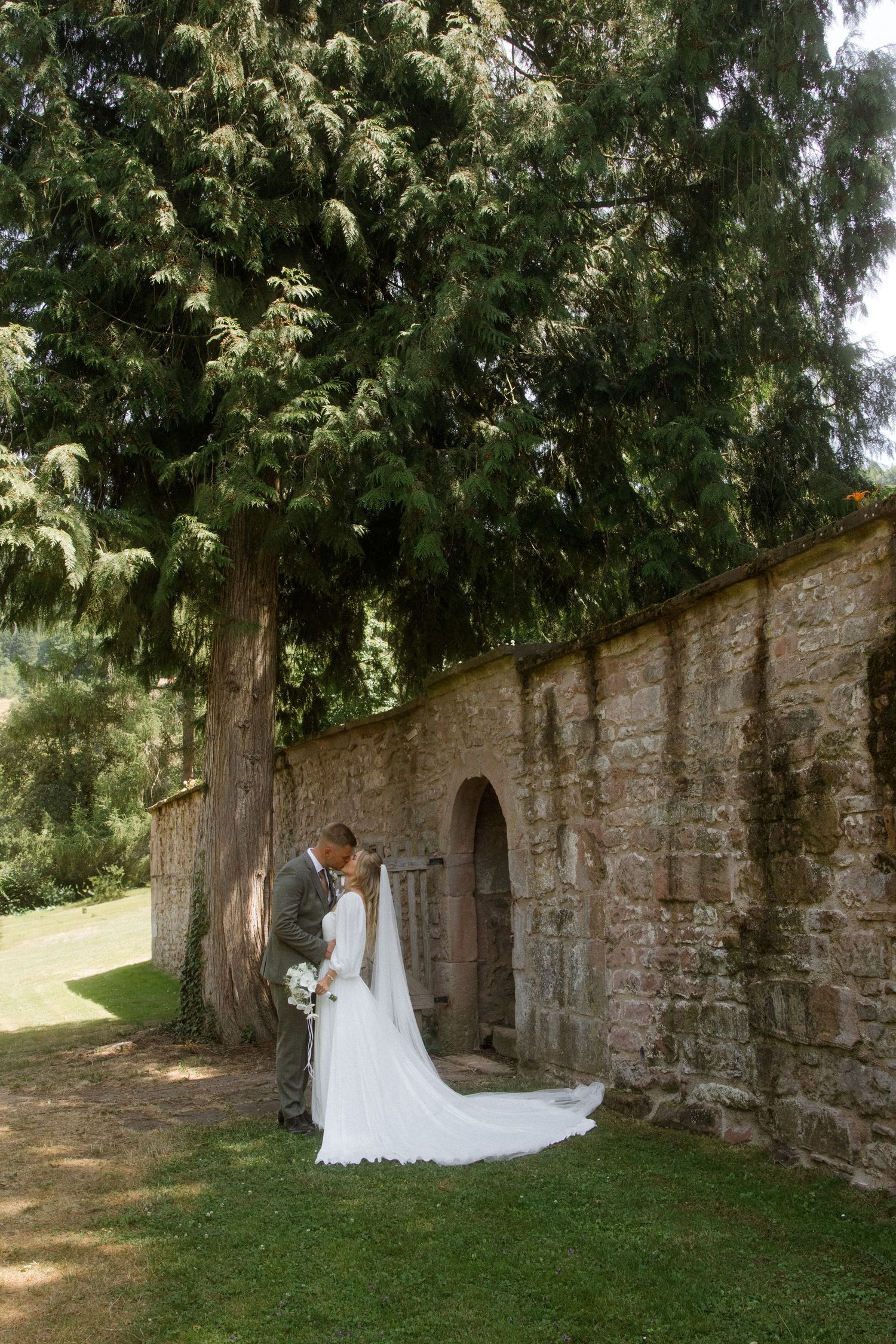 Wedding couple poses against an old stone wall beneath towering evergreen trees.