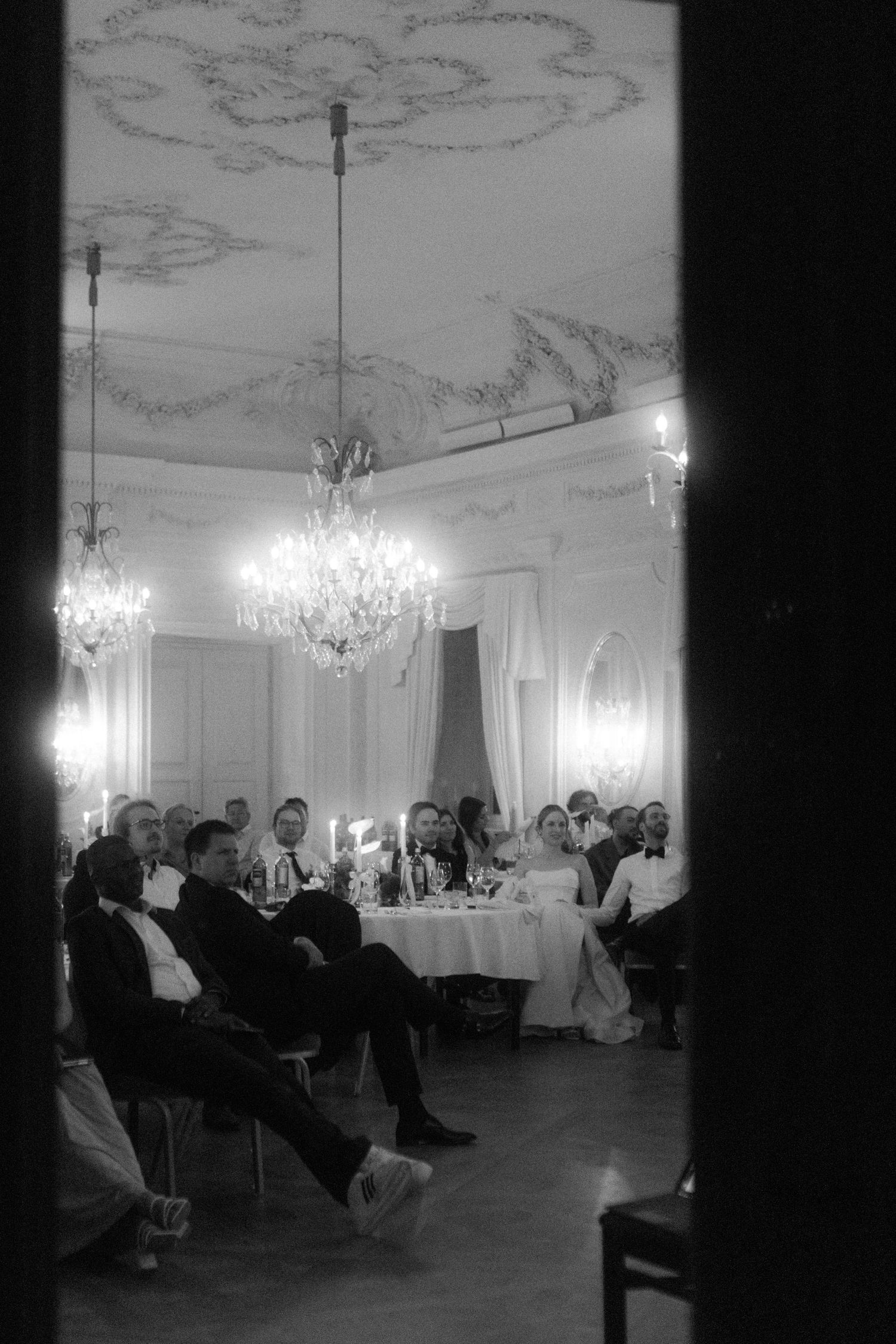 Guests seated in an elegant ballroom with ornate ceiling details and crystal chandeliers.