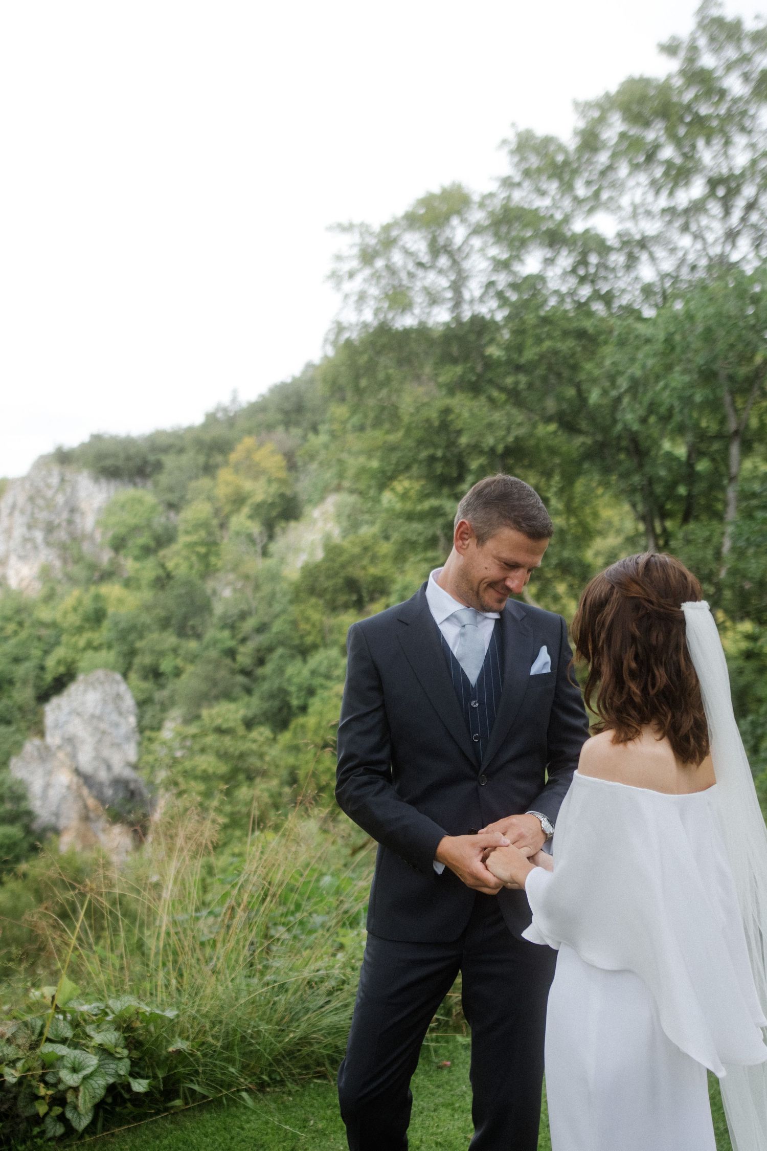 A couple holds hands during their intimate outdoor wedding ceremony against a scenic cliff backdrop.