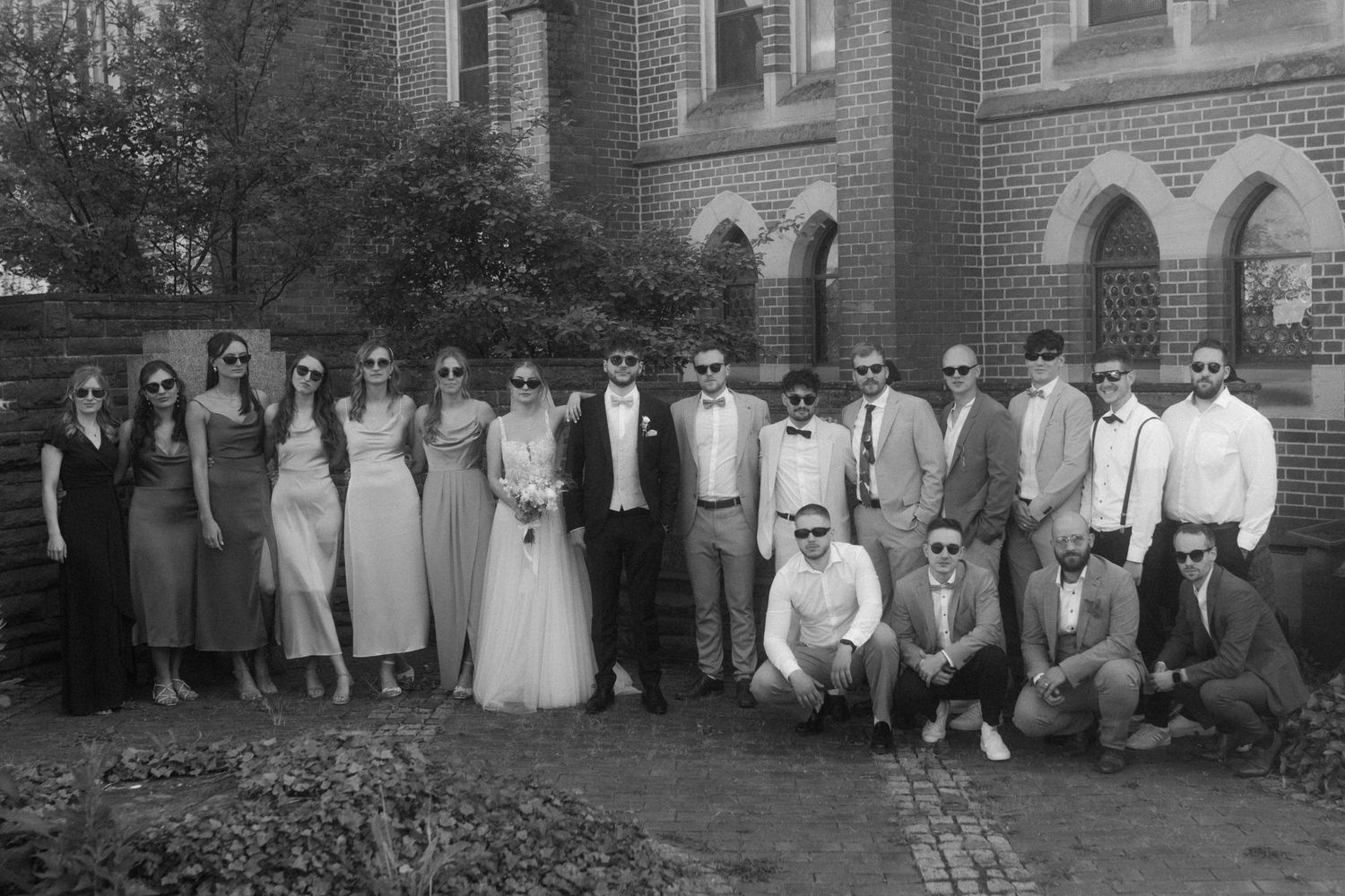 Large wedding party group photo with bridesmaids and groomsmen outside brick church.