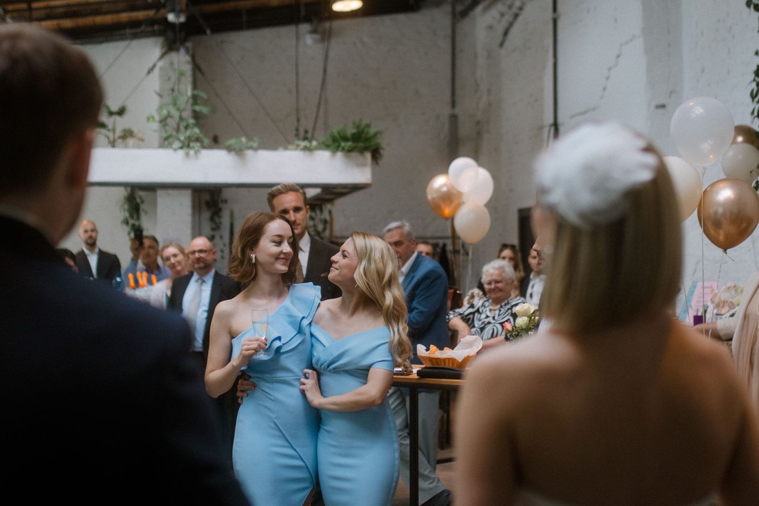 Wedding guests mingle and dance during a reception in an industrial space with white walls and hanging plants.